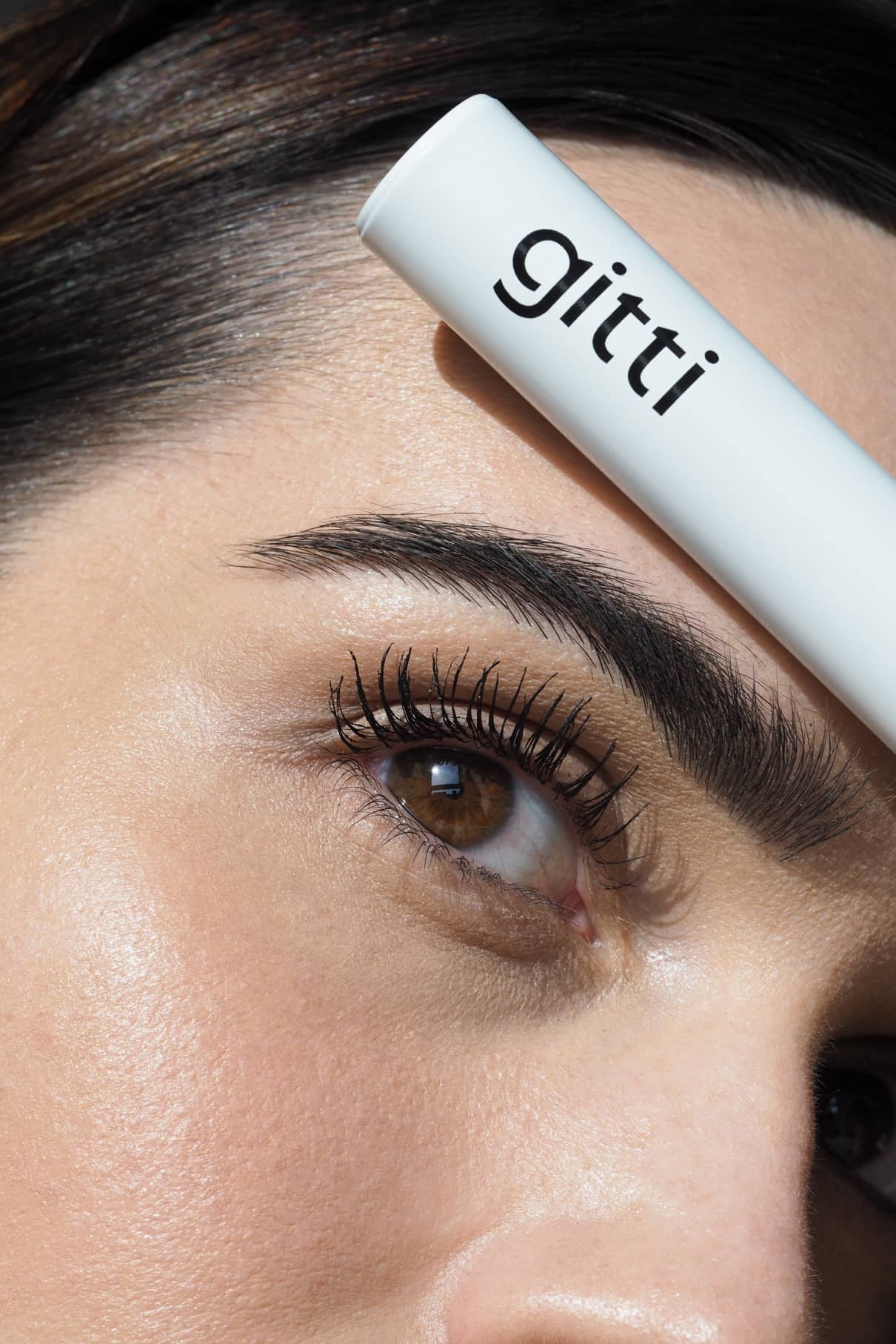 Eine Nahaufnahme des Auges einer Person mit langen, natürlichen Wimpern und einer gepflegten Augenbraue, die eine weiße Tube mit der Aufschrift Eye Level Mascara für maximales Volumen über der Braue hält.