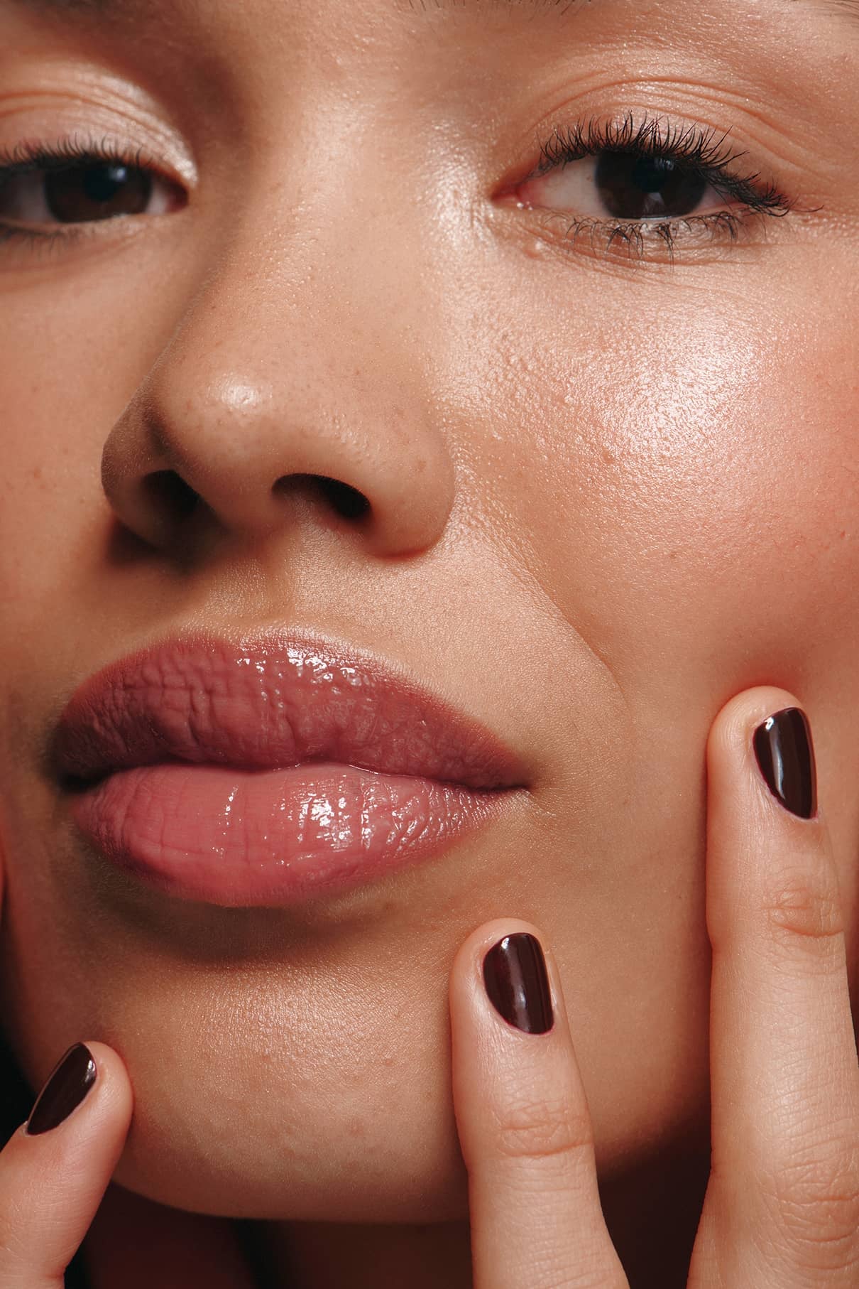 Nahaufnahme des Gesichts einer Person mit natürlichem Make-up, glänzenden Lippen mit Lip Glaze Nr. 01, und Hände mit dunklem burgunderrotem Nagellack, der die Wangen und das Kinn sanft berührt und für ein glänzendes Finish sorgt.