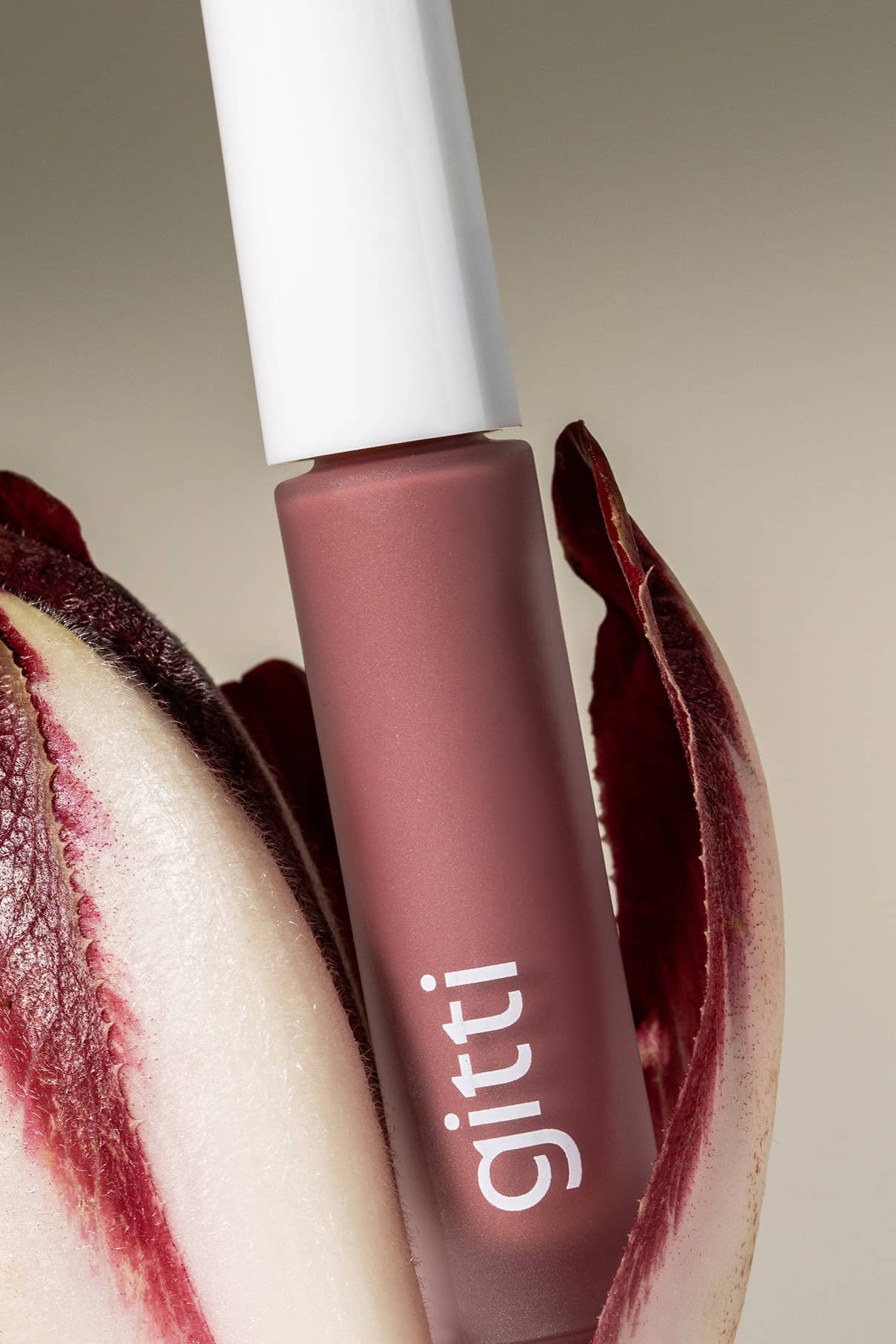 Lip Glaze no. 01, ein mauvefarbener flüssiger Lippenstift, steht aufrecht zwischen roten und weißen Endivienblättern - dieser Gloss Hybrid liefert ein hochglänzendes Finish, das auf den ersten Blick besticht.