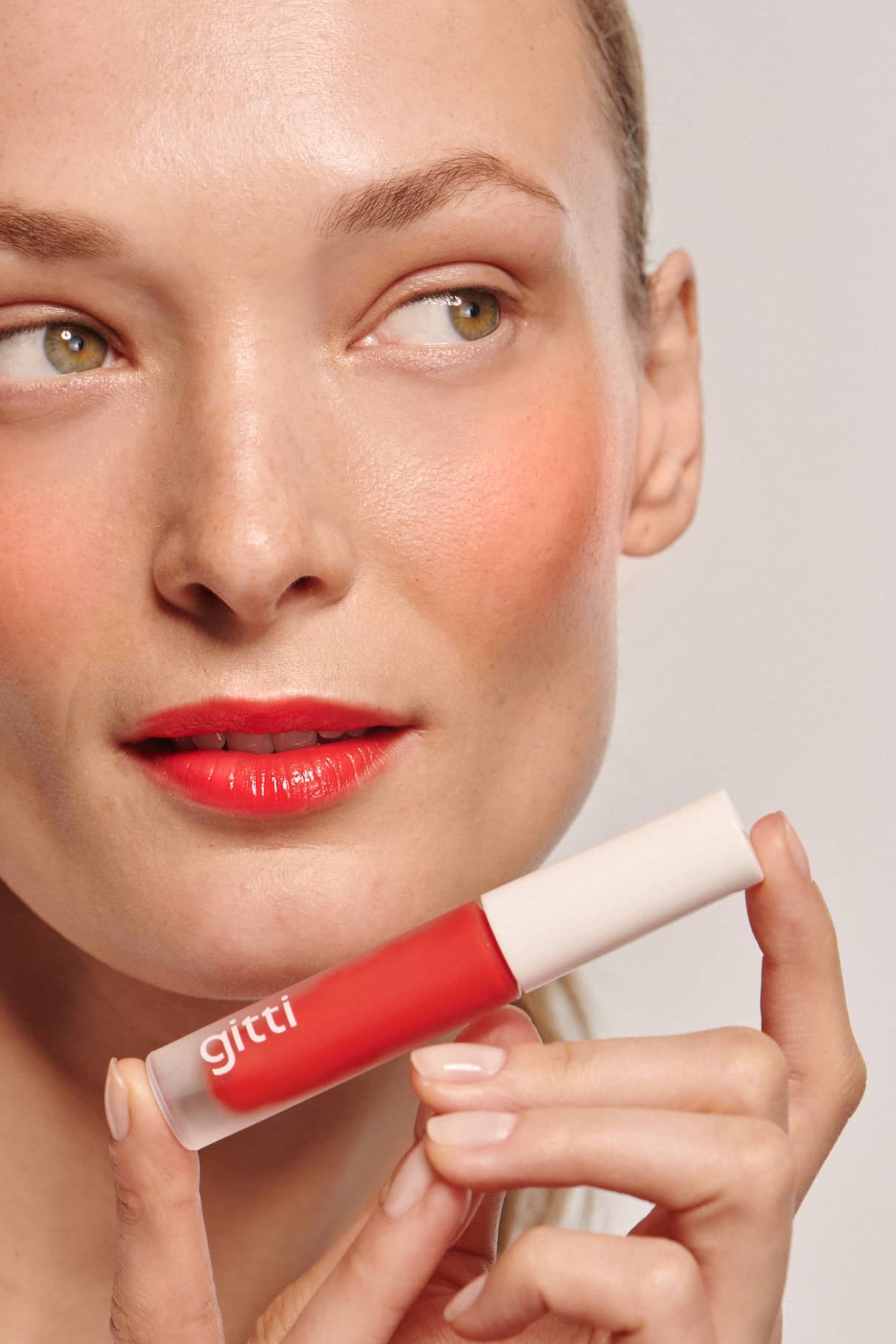 Eine Person mit heller Haut und rotem Lippenstift hält Lip Glaze no. 02 in die Nähe ihres Gesichts und präsentiert ihr hochglänzendes Finish, während sie zur Seite schaut.