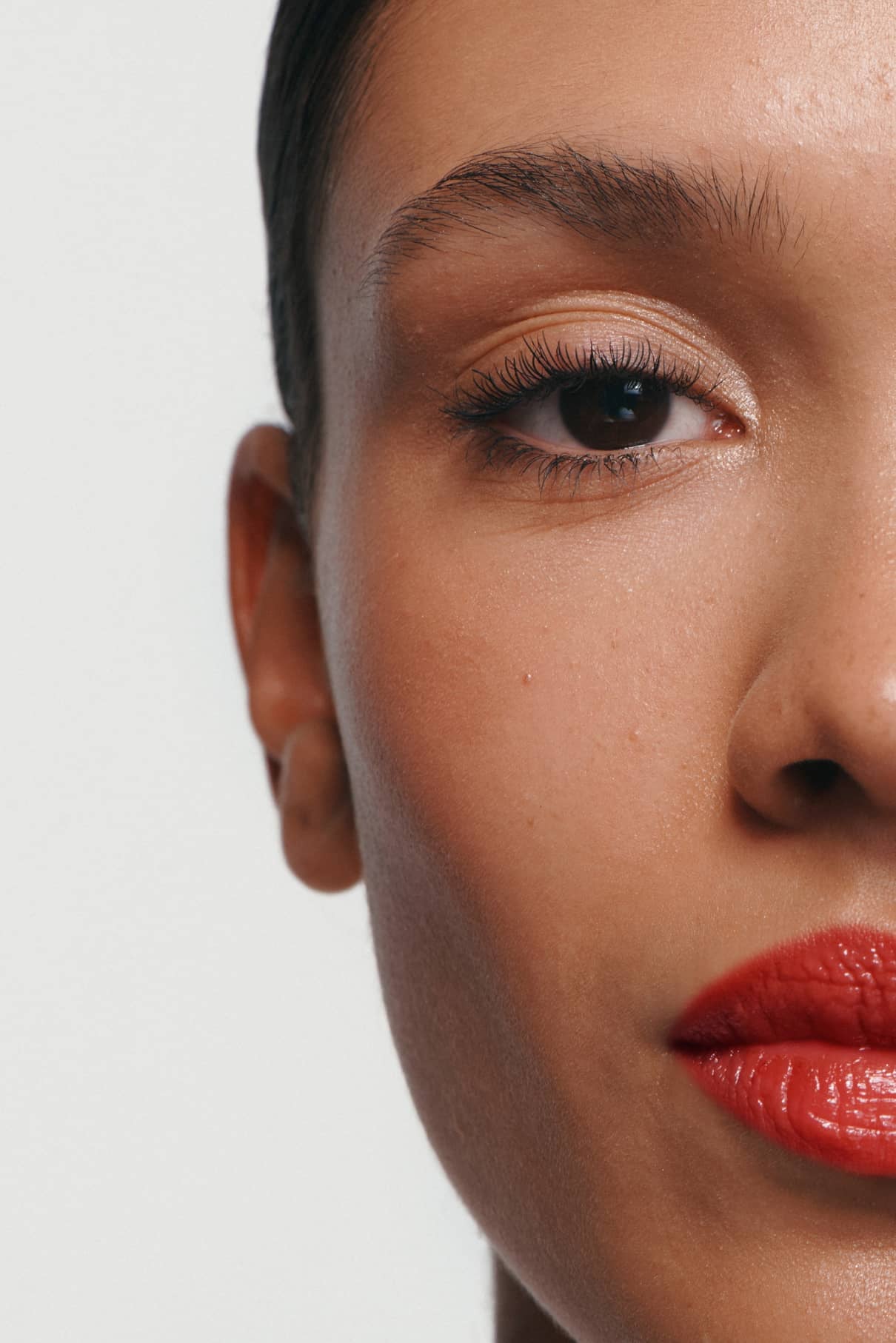 Nahaufnahme eines Frauengesichts mit glatter Haut, definierten Brauen, langen Wimpern und glänzenden roten Lippen, die mit Lip Glaze Nr. 02, vor einem schlichten Hintergrund.