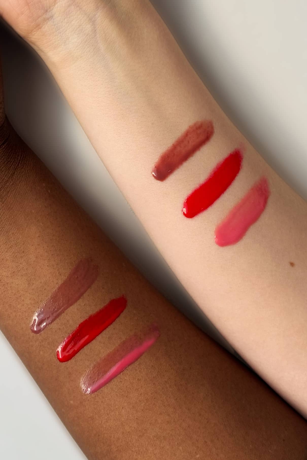 Zwei Arme mit unterschiedlichen Hauttönen zeigen Farbmuster von Lip Glaze Nr. 02 - braun, rot und pink - mit einem glänzenden Finish, das an ein luxuriöses Lippenöl erinnert, vor einem neutralen Hintergrund.