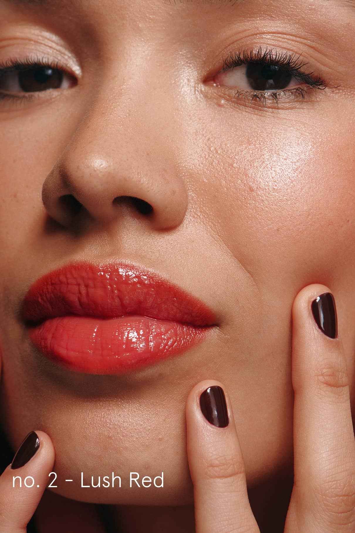 Eine Nahaufnahme zeigt eine Person mit glänzenden roten Lippen, dunklen Nägeln und taufrischer Haut, die das Signature Set trägt. Velvet Cheeks sorgen für einen sanften Schimmer. In der Ecke erscheint "No. 2 - Lush Red".