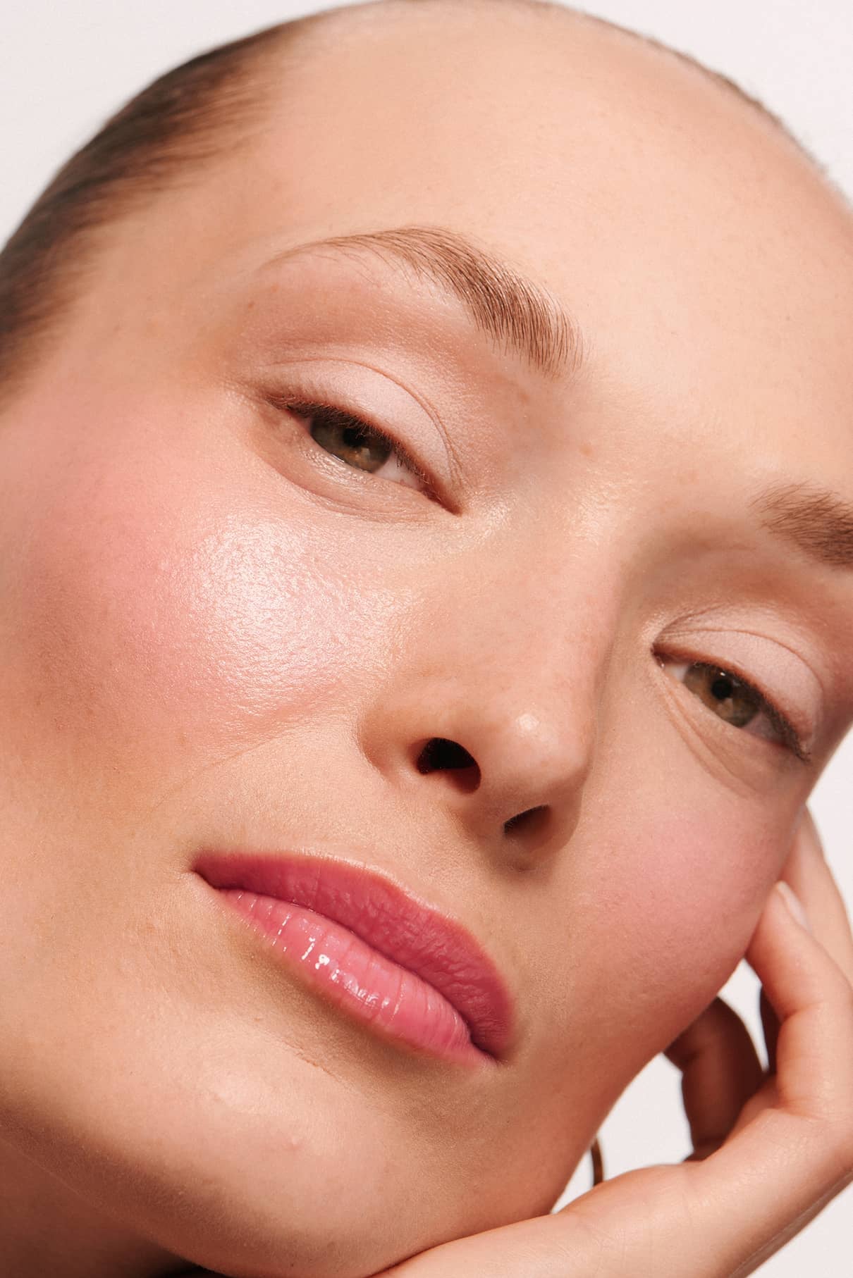 Nahaufnahme einer Person mit klarer, strahlender Haut und natürlichem Make-up, mit volleren Lippen mit hochglänzendem Finish dank Lip Glaze no. 03, während sie ihr Gesicht auf die Hand stützt, vor einem unifarbenen Hintergrund.