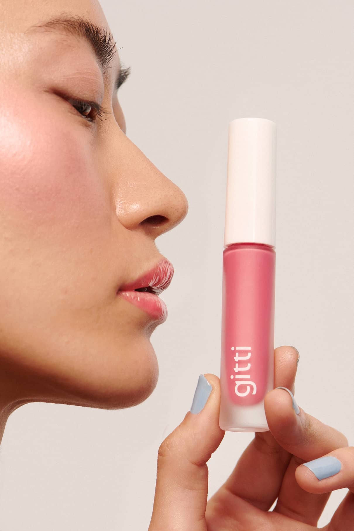 Eine Person mit rosa Make-up hält eine rosa Tube Lip Glaze No. 03 in die Nähe ihres Gesichts und präsentiert ihr hochglänzendes Finish vor einem schlichten Hintergrund.