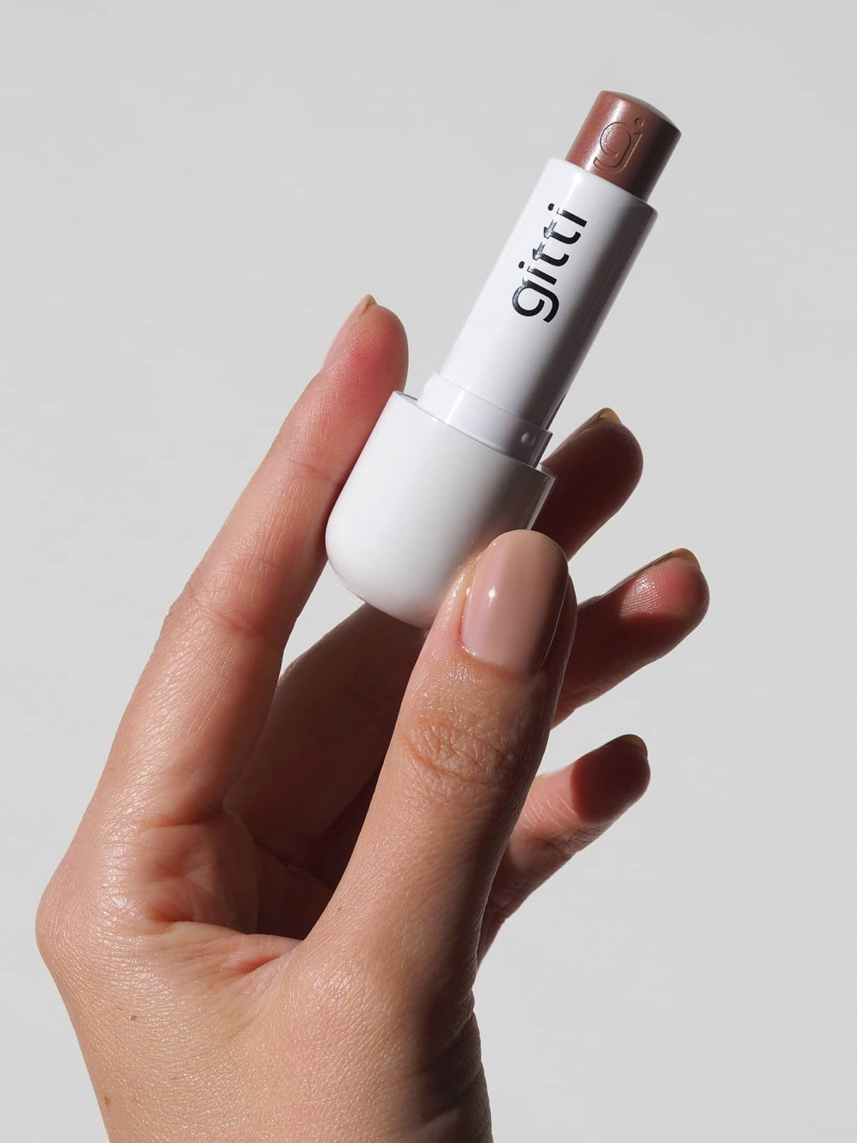 Eine Hand mit nudefarbenen Nägeln hält eine weiße Tube Lip Sync Refill no. 01, ein warmer brauner Lippenstift mit rötlichem Unterton, vor einem schlichten hellen Hintergrund.