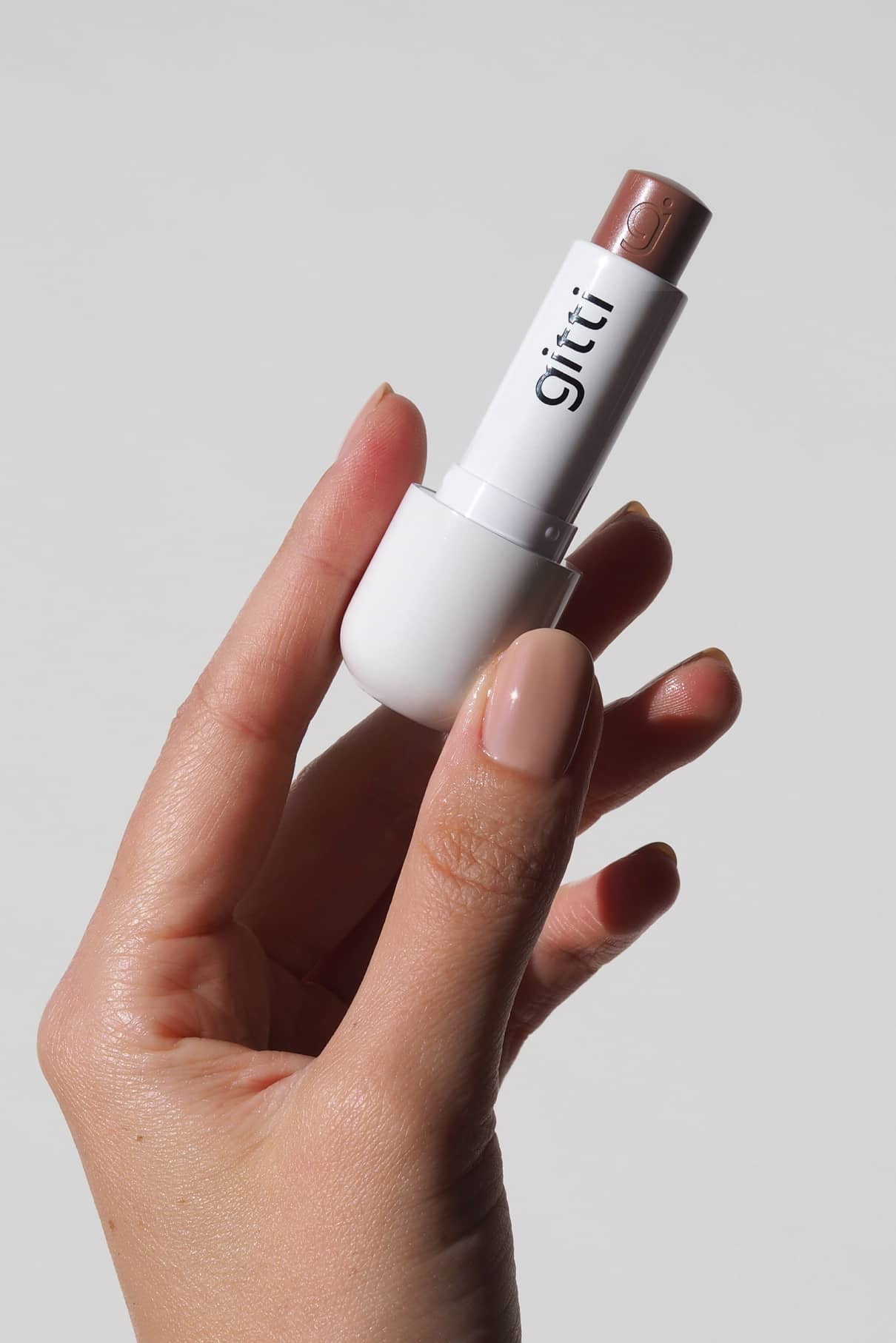 Eine Hand mit nudefarbenen Nägeln hält eine weiße Tube des gitti Celebration Trio Lippenstiftsets in einem Braunton neben dem schlanken Lip Sync Case vor einem schlichten hellen Hintergrund.