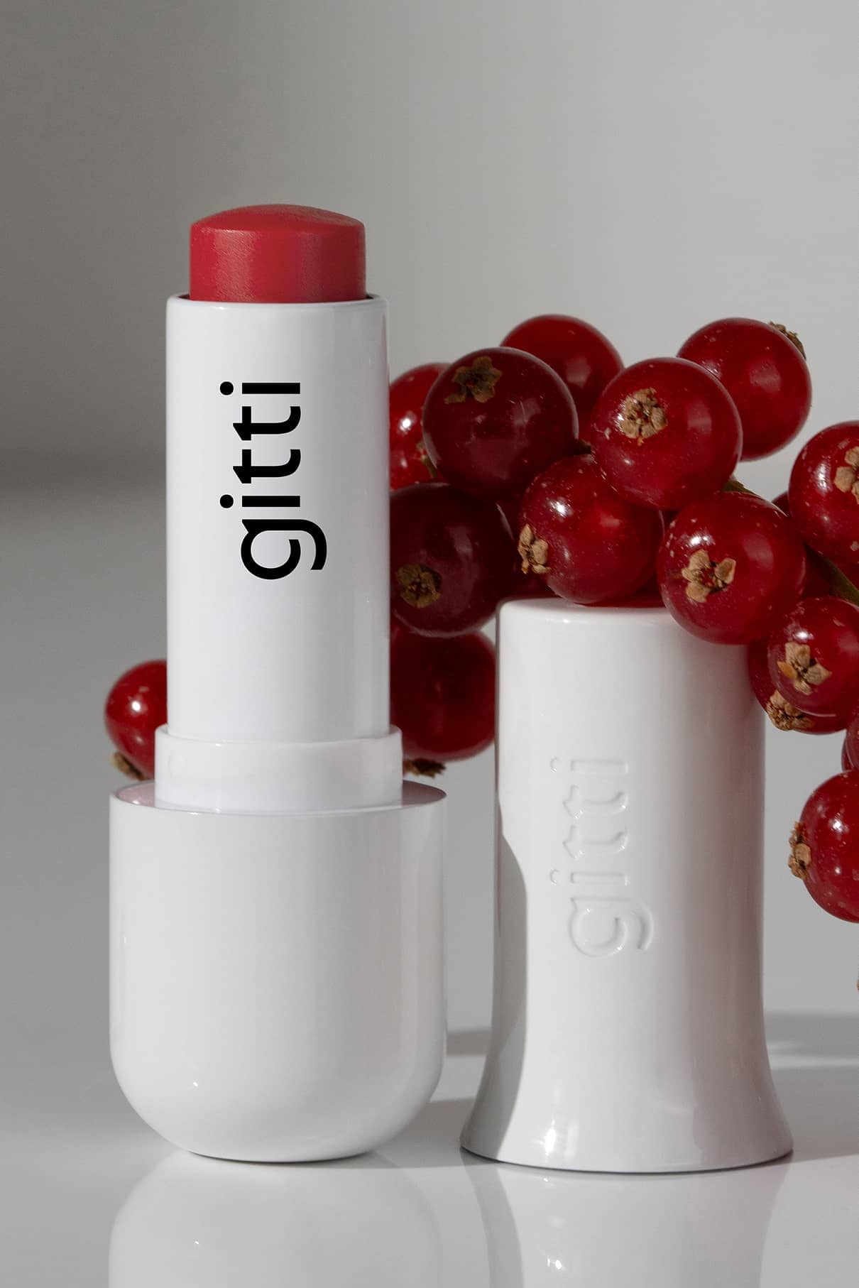 Eine geöffnete rote Lippenstifthülse mit der Aufschrift gitti steht neben ihrer Kappe und einer Gruppe roter Beeren auf einer weißen Fläche - perfekt für deine Lip Sync Momente mit dem Lip Sync Kit.