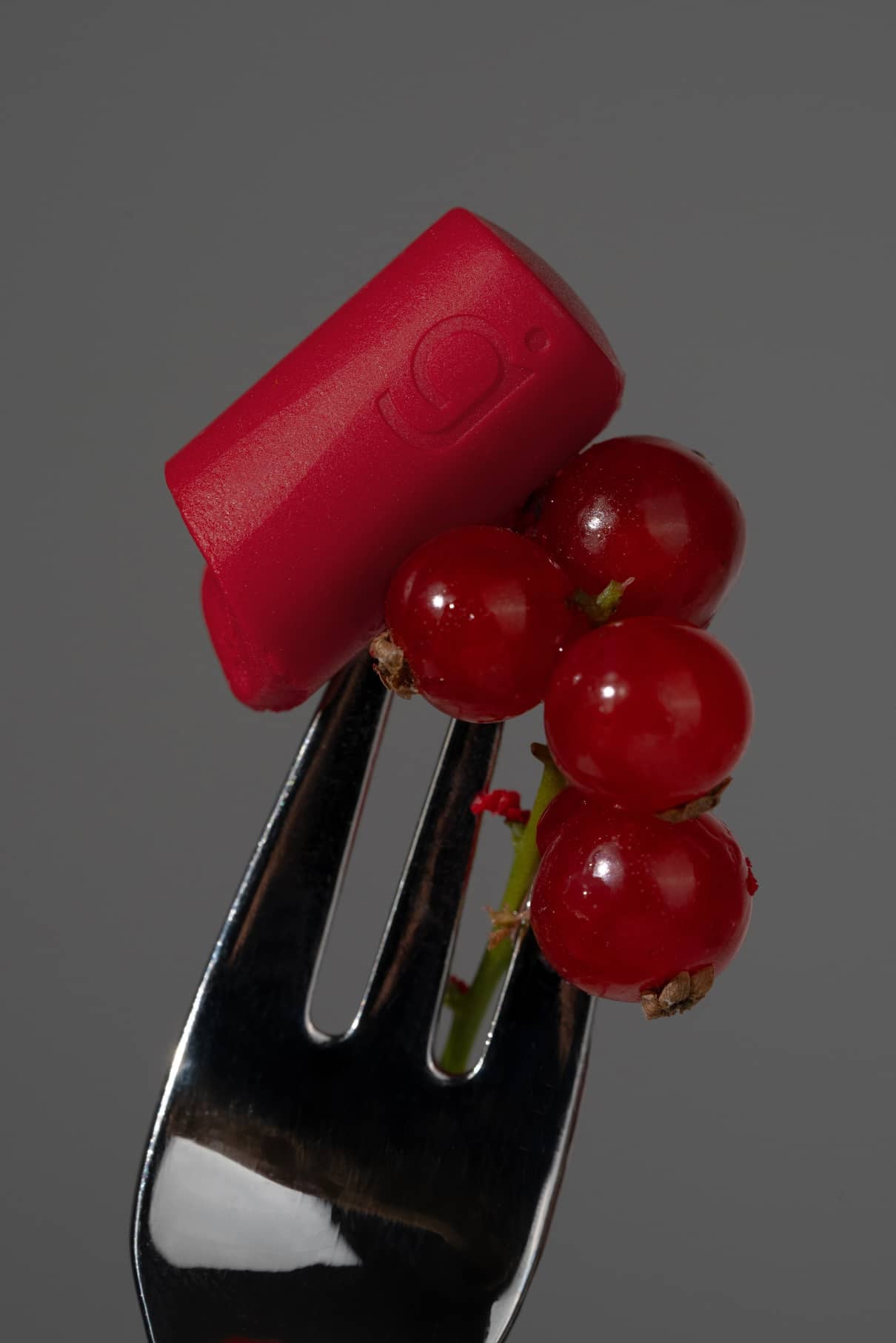 Eine Metallgabel hält ein Bündel roter Johannisbeeren und den Lippenstift Lip Sync Refill No. 02 Lippenstift, beides vor einem schlichten grauen Hintergrund.