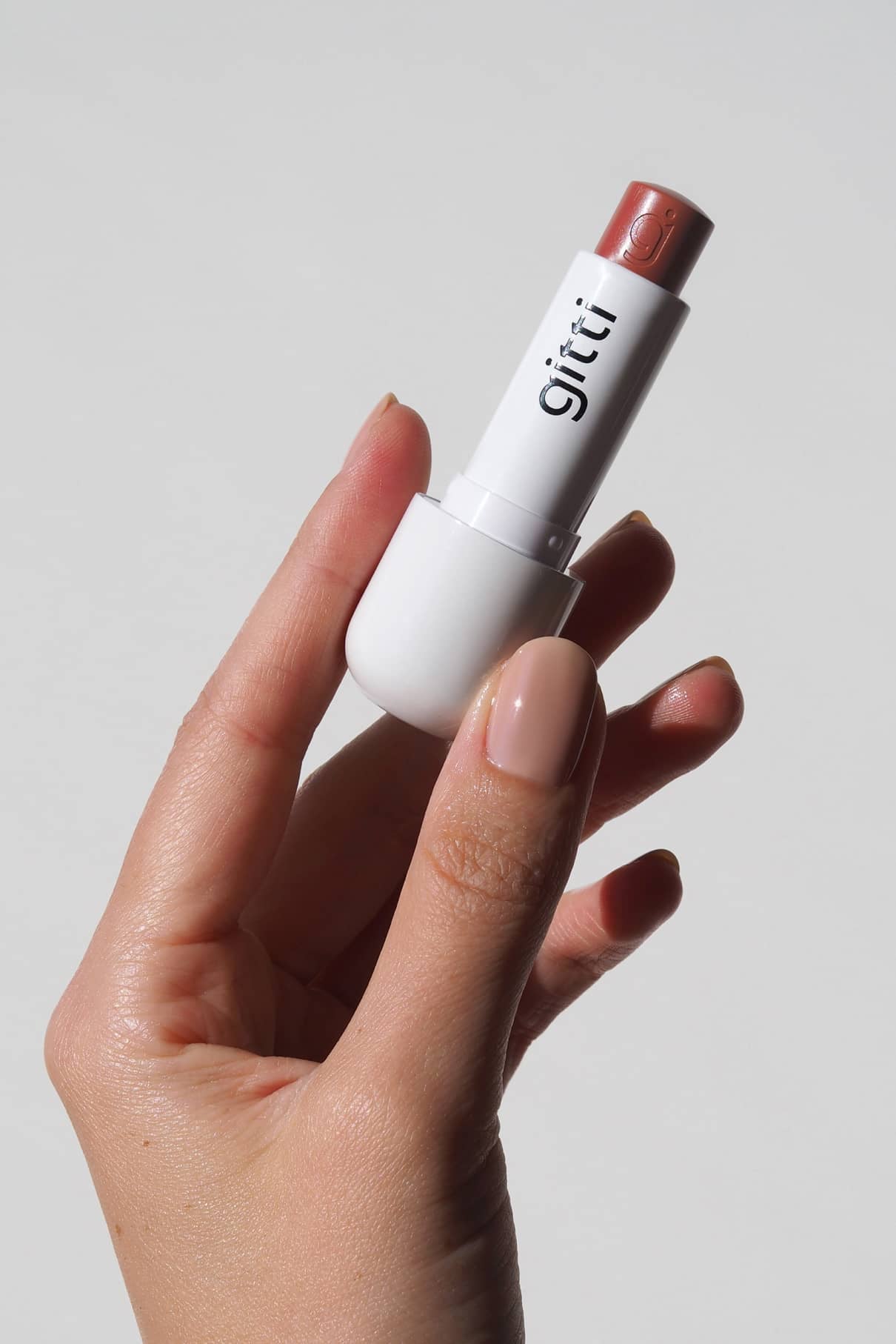 Eine Hand mit manikürten Nägeln hält Lip Sync Refill no. 03, einen nudefarbenen Gitti-Lippenstift in einer weißen Tube - perfekt für warme oder neutrale Hauttypen; dieser rosa Korall-Ton schmeichelt jedem Look vor einem schlichten Hintergrund.