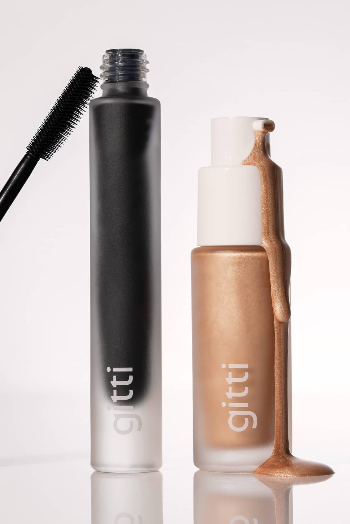 Eine schwarze Mascara-Tube mit einem Zauberstab und ein Fläschchen mit Highlighter, beide mit der Aufschrift gitti Fresh Up Set versehen, stehen auf einem weißen Hintergrund, wobei Highlighter an der Seite des Fläschchens heruntertropft.