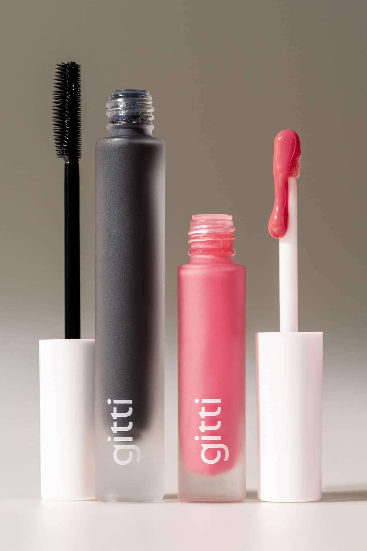 Das Lip & Lash Duo besteht aus zwei offenen Kosmetiktuben mit weißen Kappen: eine mit schwarzer Mascara und Bürste, die andere mit rosa Lip Glaze und Zauberstab. Beide sind mit gitti-ein perfektes Set für Ihre Make-up-Routine beschriftet.