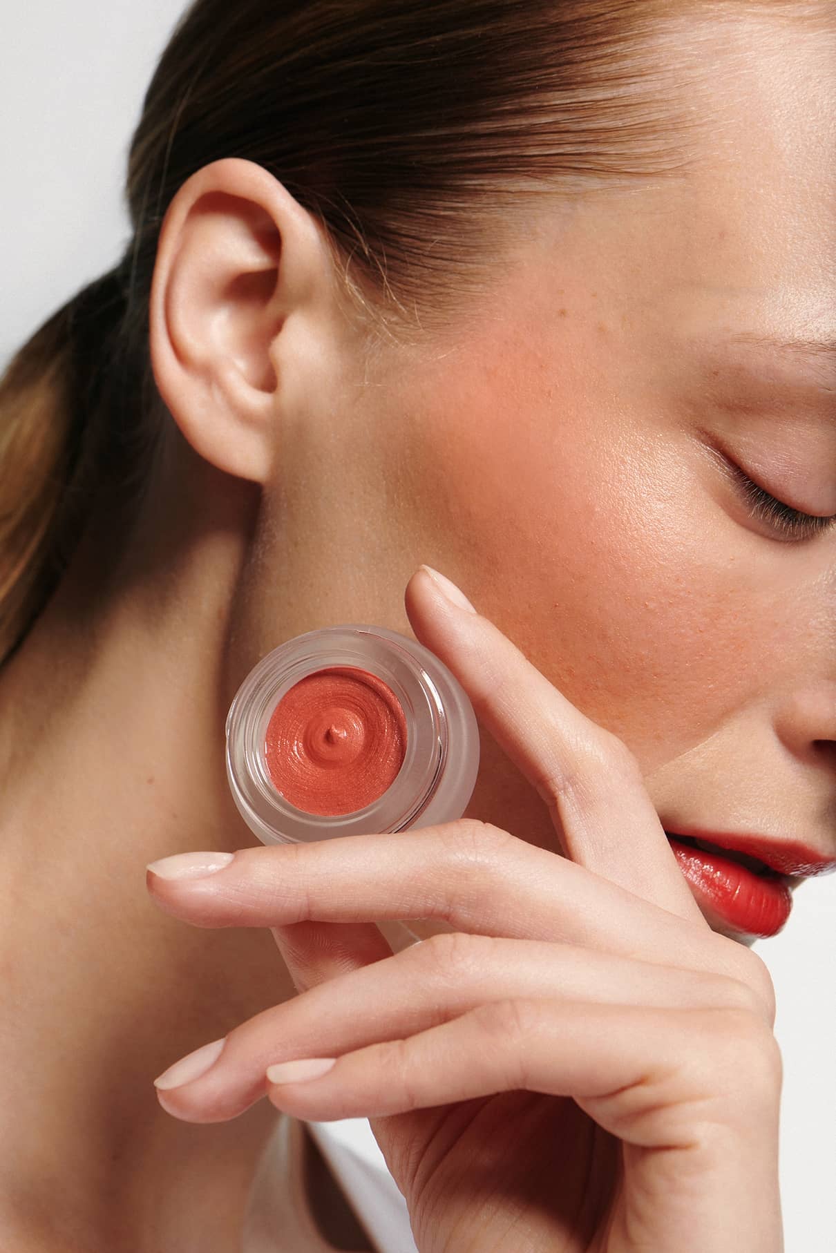 Eine Person hält eine offene Dose Velvet Cheeks no. 02 mousse-blush in Korall-Orange in der Nähe ihrer Wange, mit geschlossenen Augen und leicht geschürzten Lippen.