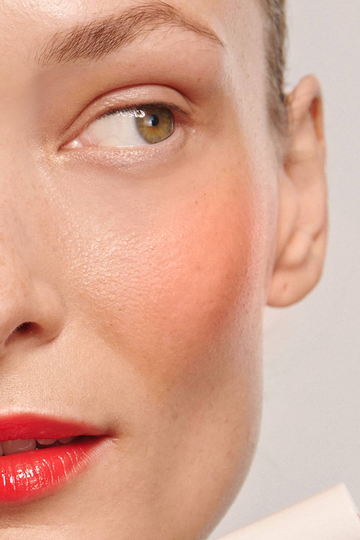 Nahaufnahme des Gesichts einer Frau, die ihre grünen Augen, Augenbrauen, Ohren und roten Lippen mit natürlicher Hauttextur und Velvet Cheeks no. 02 mousse-blush in Korall-Orange-perfekt verblendet für ein silikonfreies Finish.