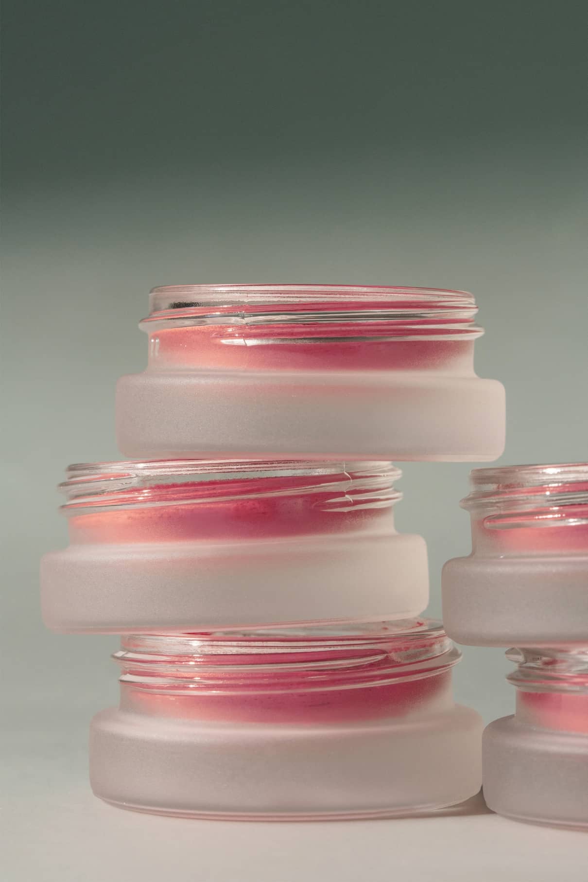 Vier Milchglasdosen mit Velvet Cheeks Nr. 03 Mousse-Blush in Pastell-Rosa sind vor einem hellgrünen Hintergrund gestapelt. Die teilweise geöffneten Gläser geben den Blick auf die silikonfreie Creme im Inneren frei.