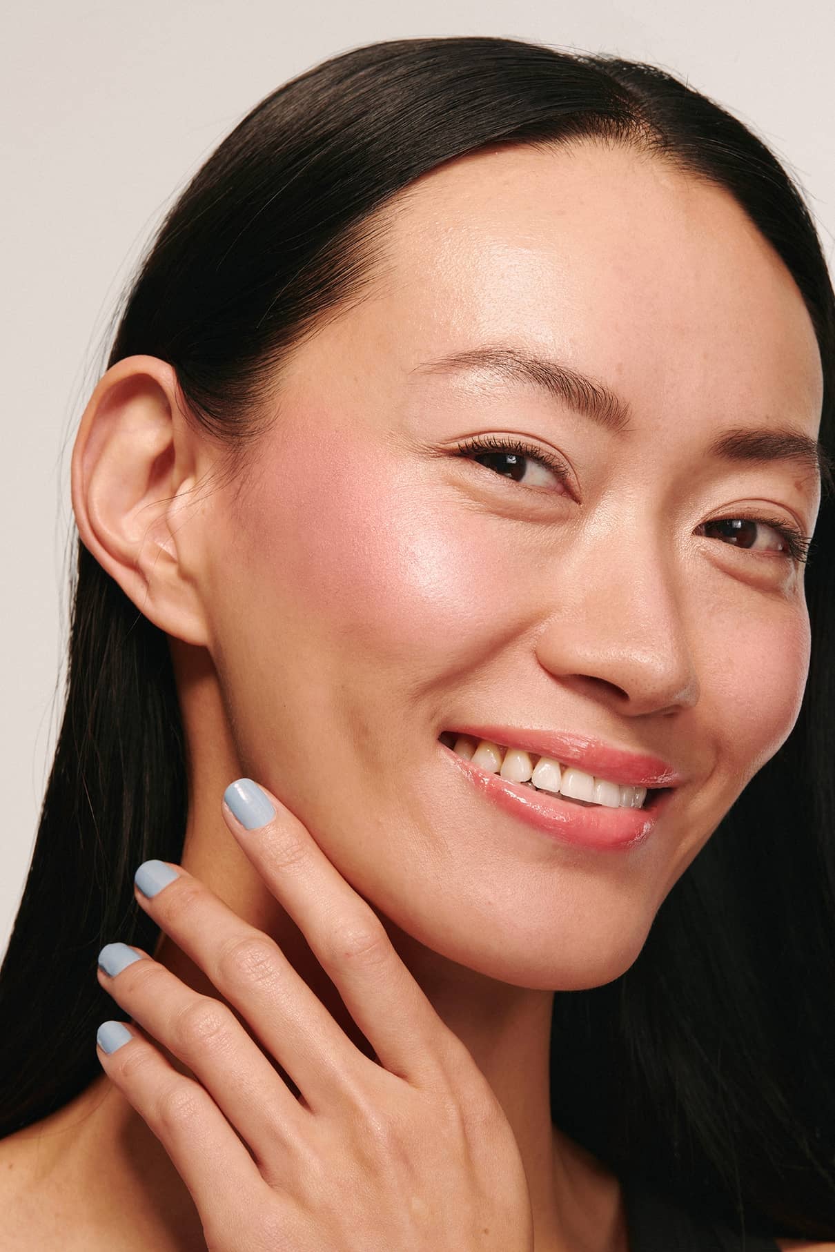 Eine Frau mit langen schwarzen Haaren lächelt, ihre helle Haut strahlt unter den Velvet Cheeks no. 03 Mousse-Blush Highlights. Sie trägt hellblauen Nagellack und berührt ihr Gesicht, wobei sie das silikonfreie Rouge für einen makellosen, natürlichen Look zur Geltung bringt.