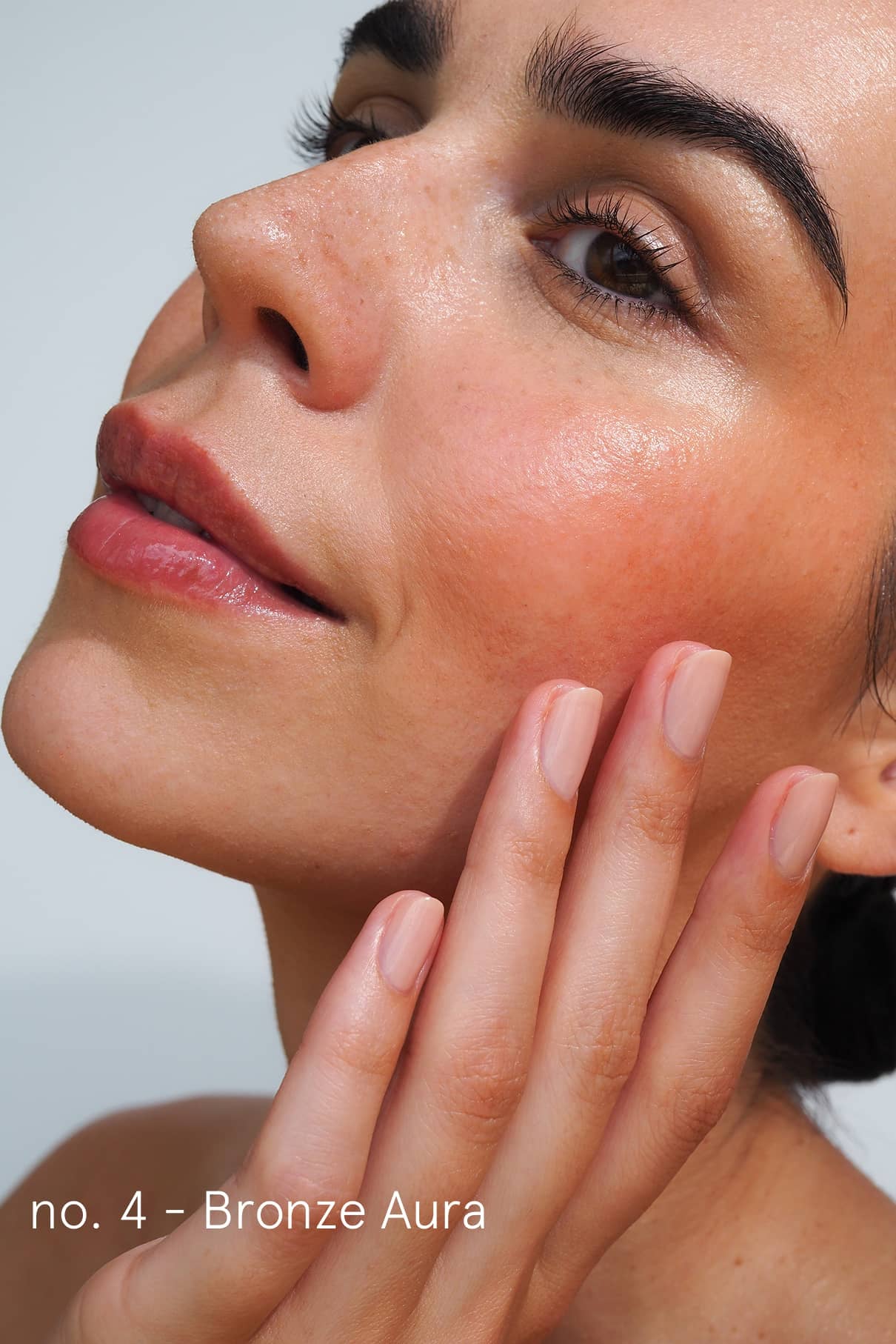 Nahaufnahme einer Person mit strahlender Haut, natürlichem Make-up und manikürten Nägeln, die durch das Summer Glow Set verbessert wurden, die ihr Gesicht berührt. Der Text auf dem Bild lautet: no. 4 - Bronze Aura.
