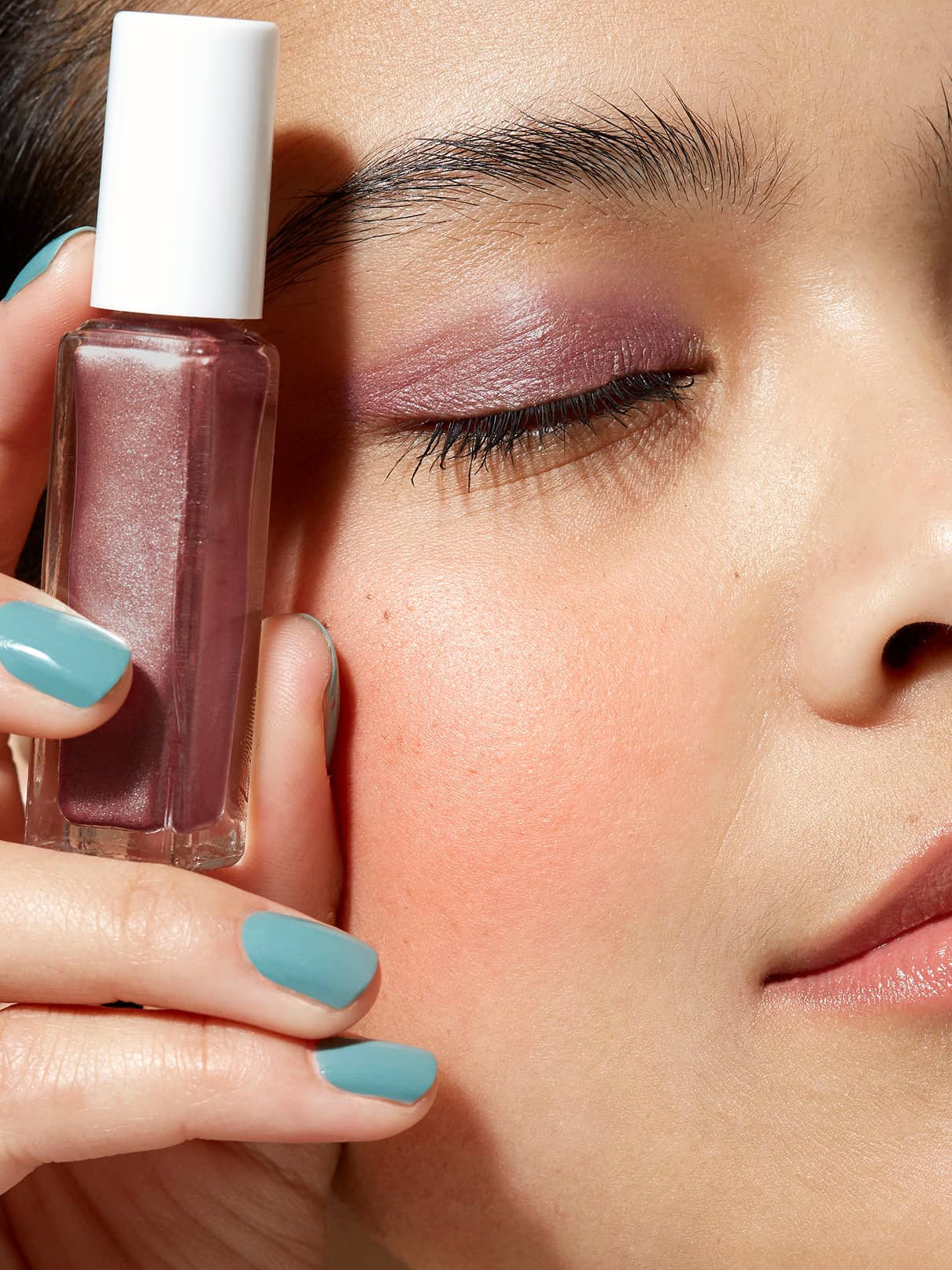 Nahaufnahme einer Person mit Beerenton-Lidschatten und tealfarbenen Nägeln, die Visionist Nr. 01 mauve polish in der Nähe ihres Gesichts hält, ein Auge geschlossen - ein cooler Look mit kühler Unterton.