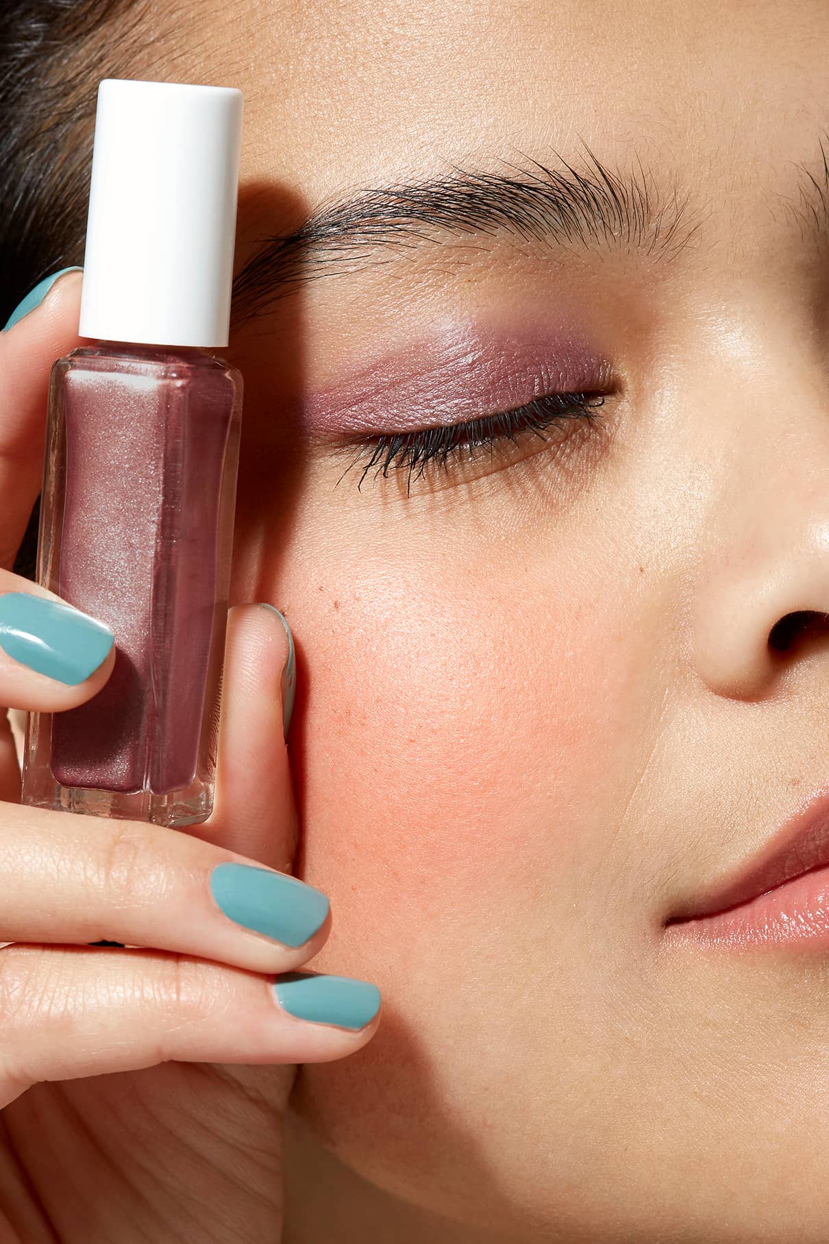 Nahaufnahme einer Person mit Beerenton-Lidschatten und tealfarbenen Nägeln, die Visionist Nr. 01 mauve polish in der Nähe ihres Gesichts hält, ein Auge geschlossen - ein cooler Look mit kühler Unterton.
