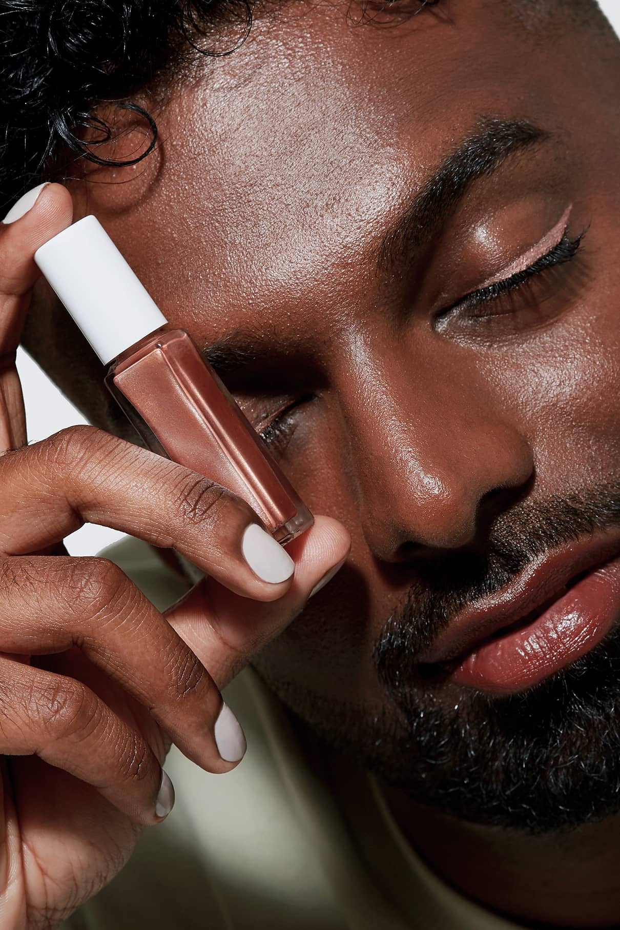 Mit geschlossenen Augen hält eine Person Visionist no. 02, ein schimmerndes, kupferfarbenes Liquid-Makeup, nahe ans Gesicht und zeigt dazu passenden Eyeliner und gepflegtes Gesichtshaar - ideal für einen neutralen Look.