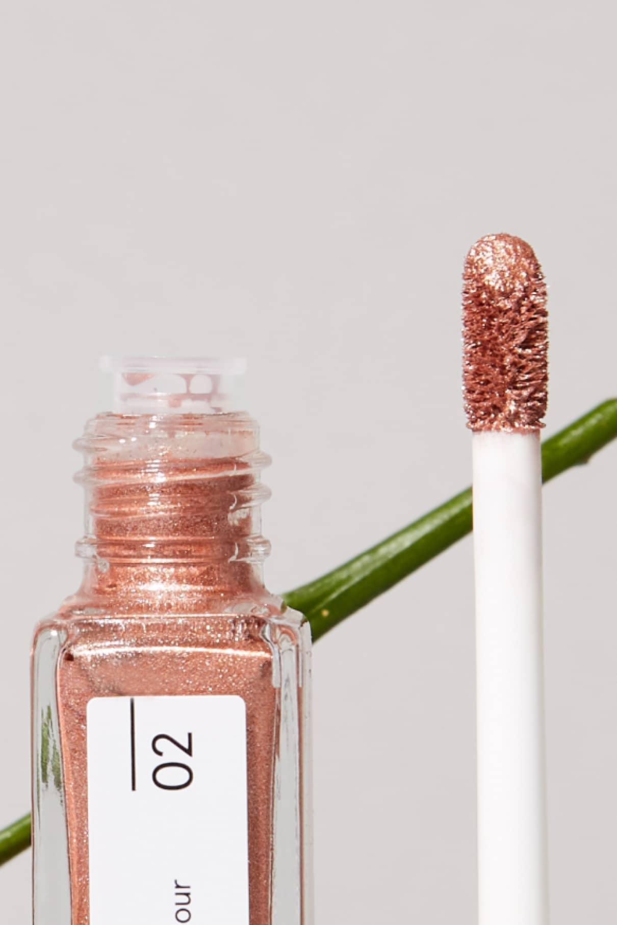 Eine Nahaufnahme von Visionist Nr. 02, einem flüssigen Lidschatten in schimmerndem Roségold mit neutralem Unterton in einer durchsichtigen Tube mit der Aufschrift 02, daneben der Applikatorstab und im Hintergrund ein grüner Pflanzenstängel.