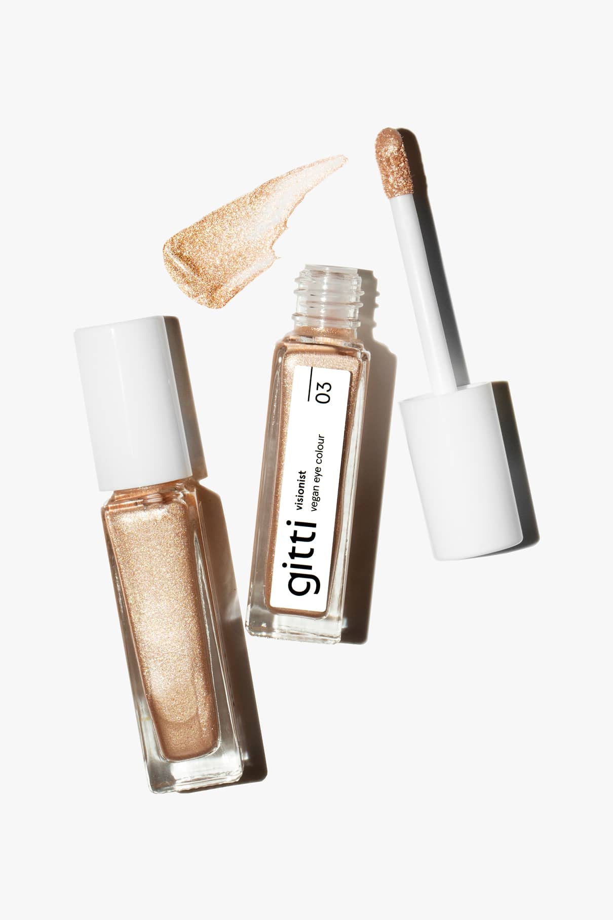 Zwei offene Tuben des schimmernden Flüssiglidschattens Visionist Nr. 03 mit weißer Kappe, eine davon mit abgenommenem Applikator, und ein Stück schimmerndes Gold - perfekt, um jedem Look einen Hauch von Glamour zu verleihen.