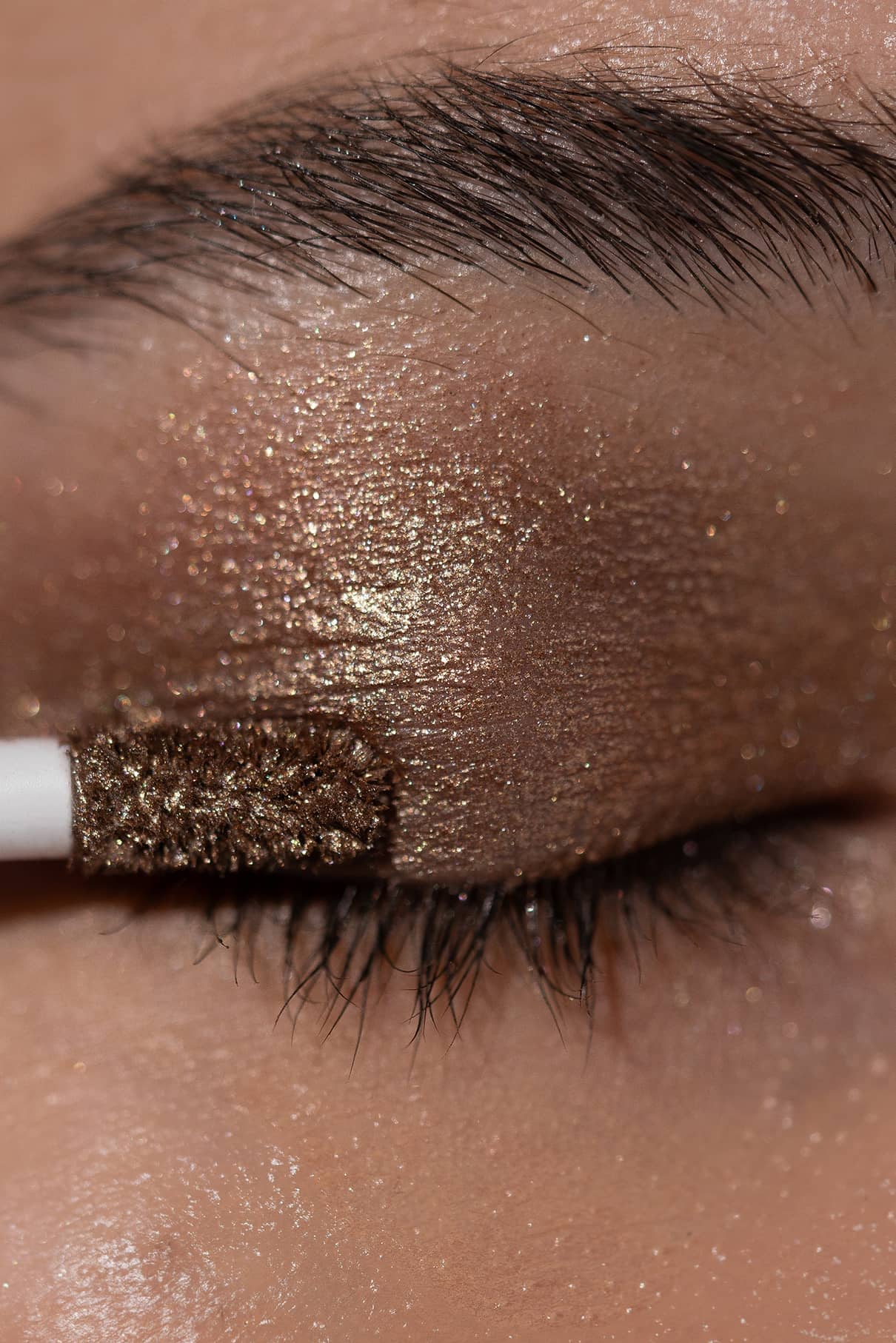 Nahaufnahme eines geschlossenen Auges, während Visionist Nr. 04, ein glitzernder, langanhaltender Lidschatten in warmen Gold- und Sienna Glow-Tönen, mit einem Applikatorstab auf das Augenlid aufgetragen wird.