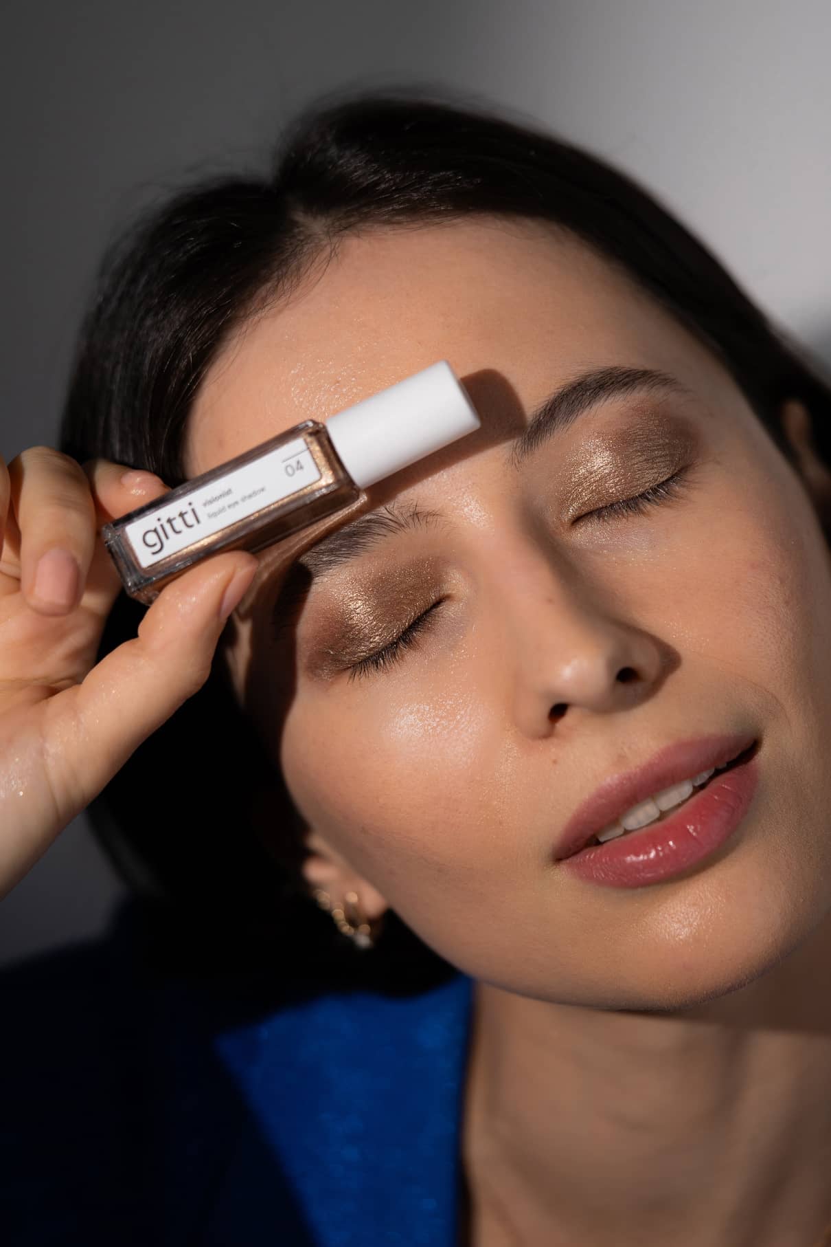 Eine Frau mit geschlossenen Augen hält sich ein kleines Fläschchen mit der Aufschrift Visionist Nr. 04 an die Stirn und betont damit Langanhaltender Lidschatten in Sienna Glow und natürlichen Lippenstift.
