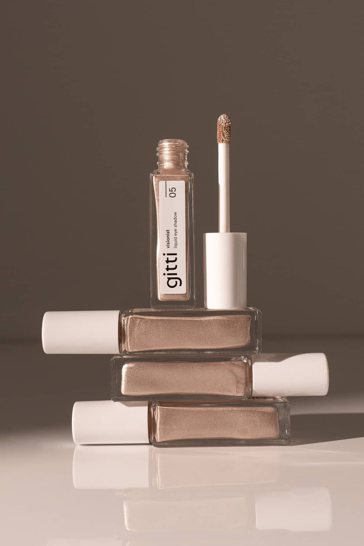 Vier rechteckige Flaschen mit beigefarbenem Metallic-Nagellack sind übereinander gestapelt, wobei die oberste geöffnet ist und ein Pinsel darin ruht. Auf dem Etikett steht "Visionist no. 05". Das schimmernde Finish erinnert an Silver Sheen oder silbernen Lidschatten.