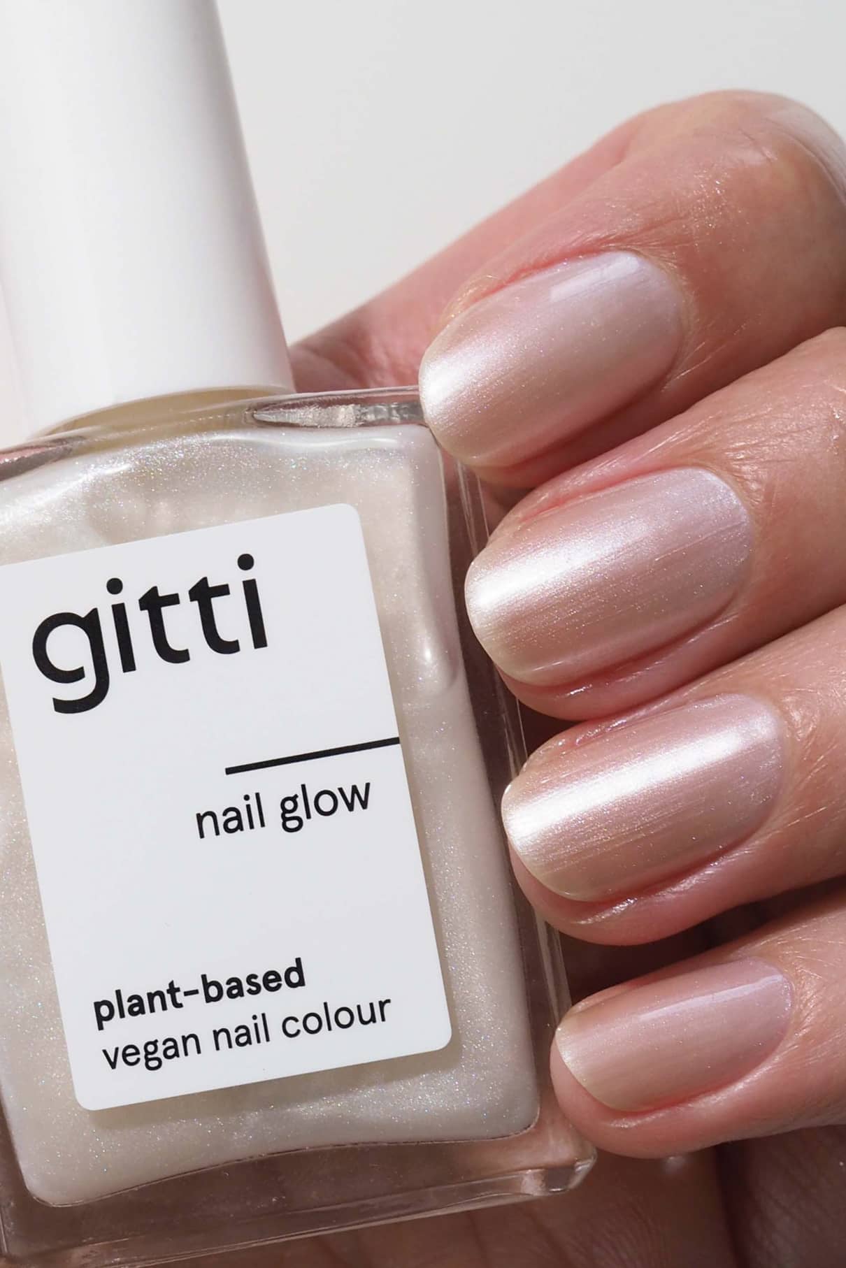 Eine Hand mit sauber manikürten Nägeln in einem perlmuttfarbenen Hellrosa hält eine Flasche gitti Nail Glow, einen veganen Nagellack auf Pflanzenbasis, der mit einem schimmernden Top Coat für zusätzlichen Glanz sorgt.