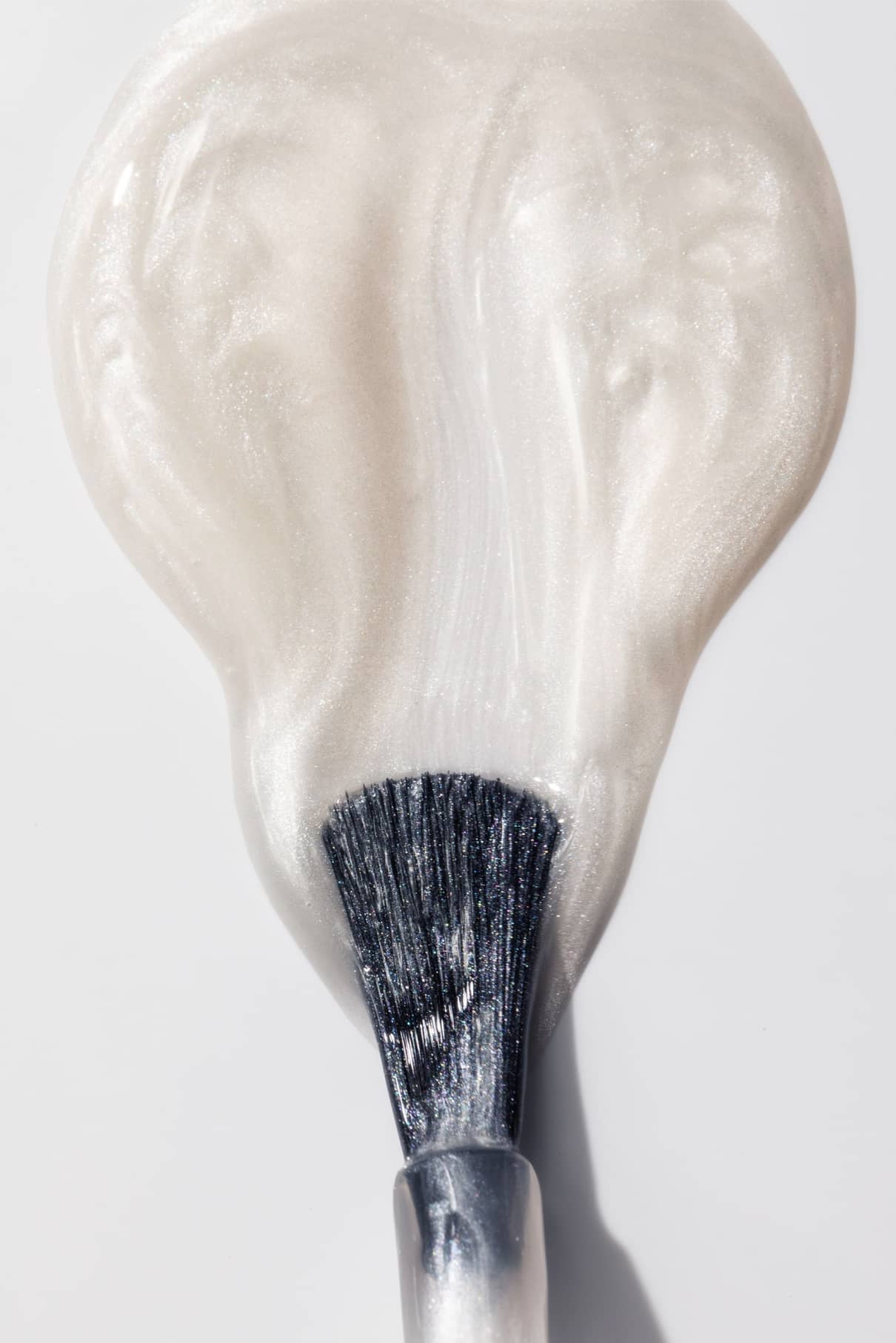 Eine Nahaufnahme zeigt einen Schminkpinsel, der Nail Glow, eine perlweiße Flüssigkeit mit schimmerndem Chromeffekt, auf einer weißen Fläche verteilt.