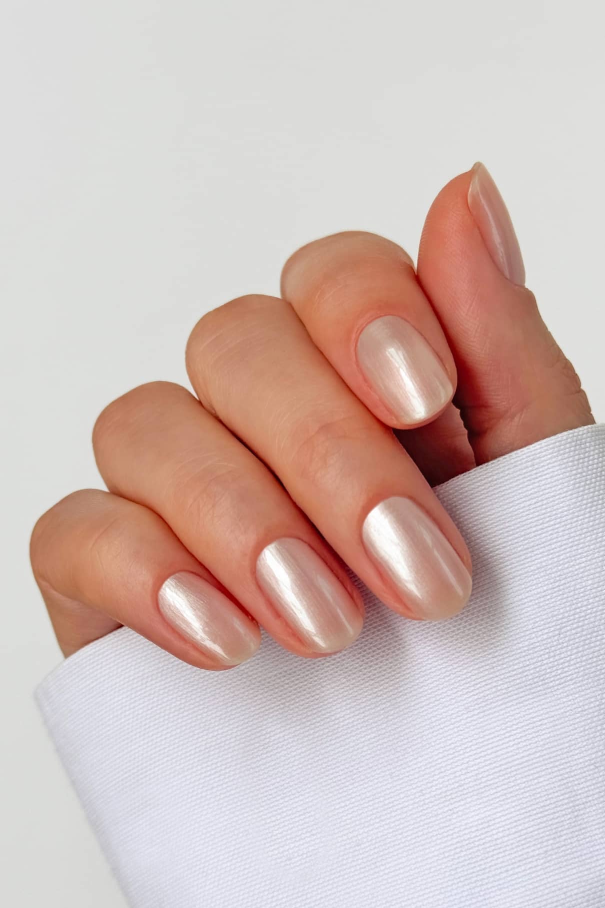 Eine Hand mit sauber manikürten Nägeln, lackiert in Nail Glow, einem perlmuttartig schimmernden, hellrosa Farbton mit glänzender Oberfläche, ruht vor einem weißen Stoffhintergrund.