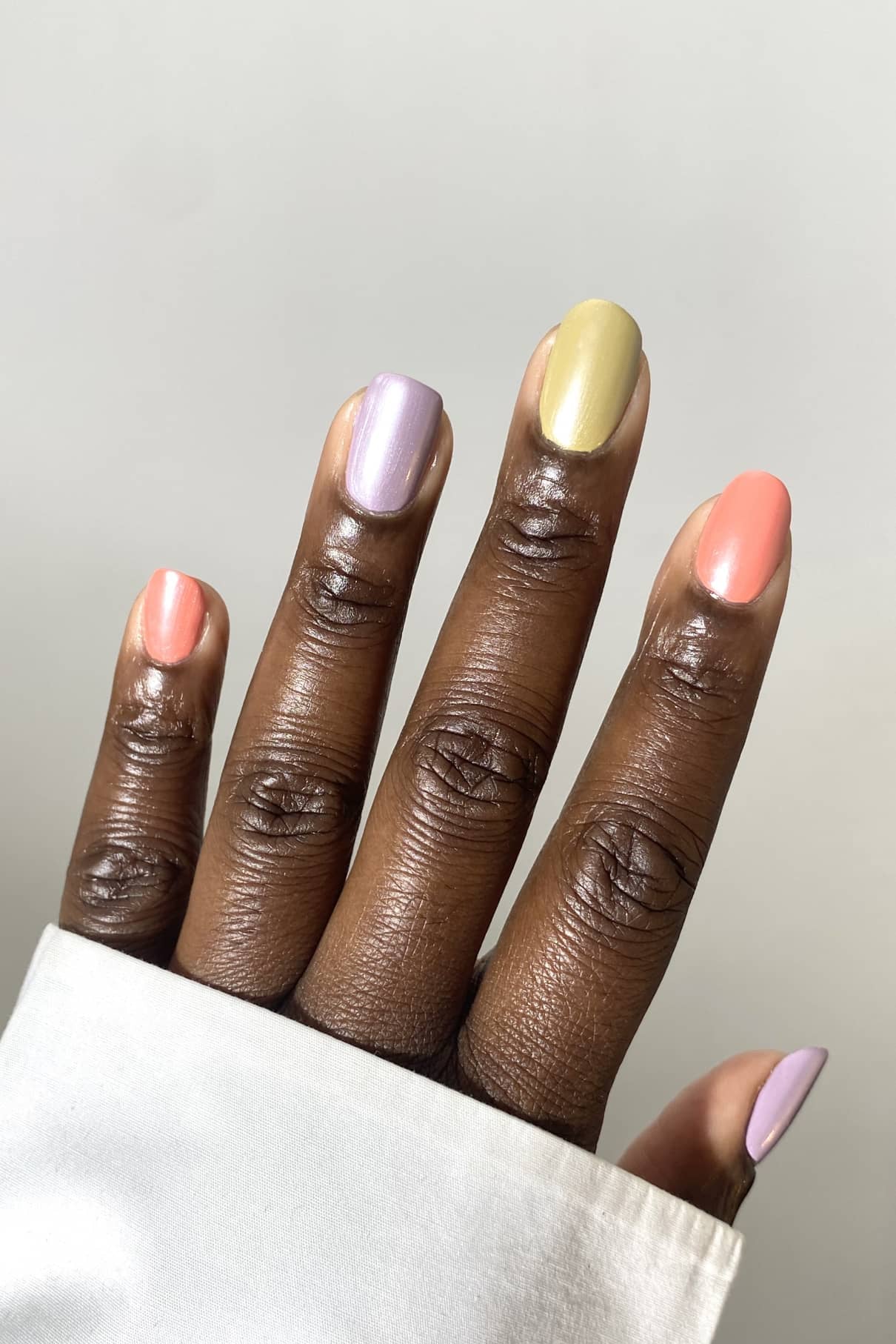 Eine Hand mit sauber manikürten Nägeln, die jeweils in korallenroten, lavendelfarbenen, gelben, rosafarbenen und violetten Pastelltönen lackiert sind - mit Nail Glow Top Coat für einen dezenten Glanz veredelt - hebt sich von einem schlichten hellen Hintergrund ab.