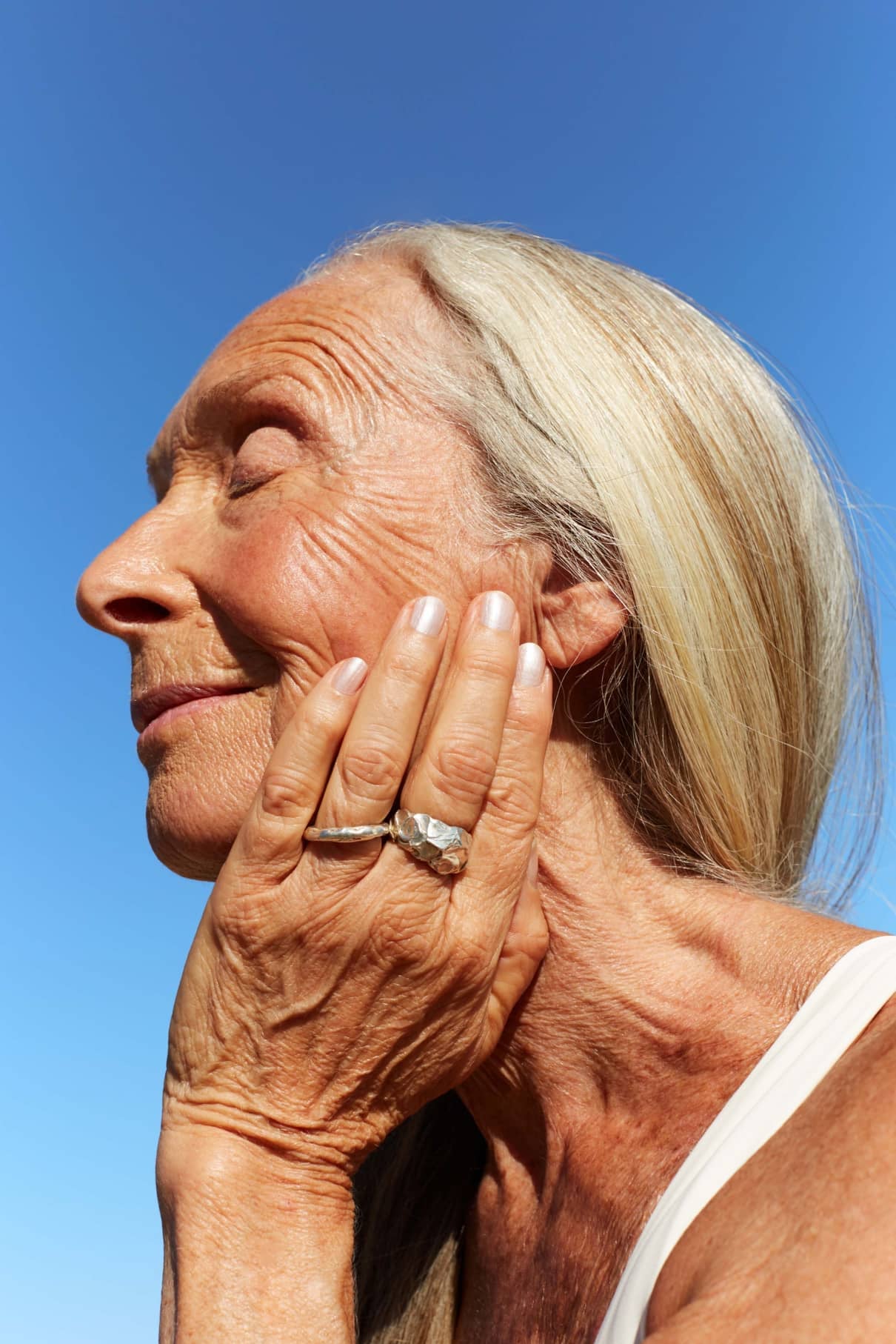 Eine ältere Frau mit langen grauen Haaren berührt sanft ihr Gesicht, die Augen geschlossen, vor einem klaren blauen Himmel. Ihre Haut leuchtet mit dem subtilen, schimmernden Chrom-Effekt von Nail Glow.