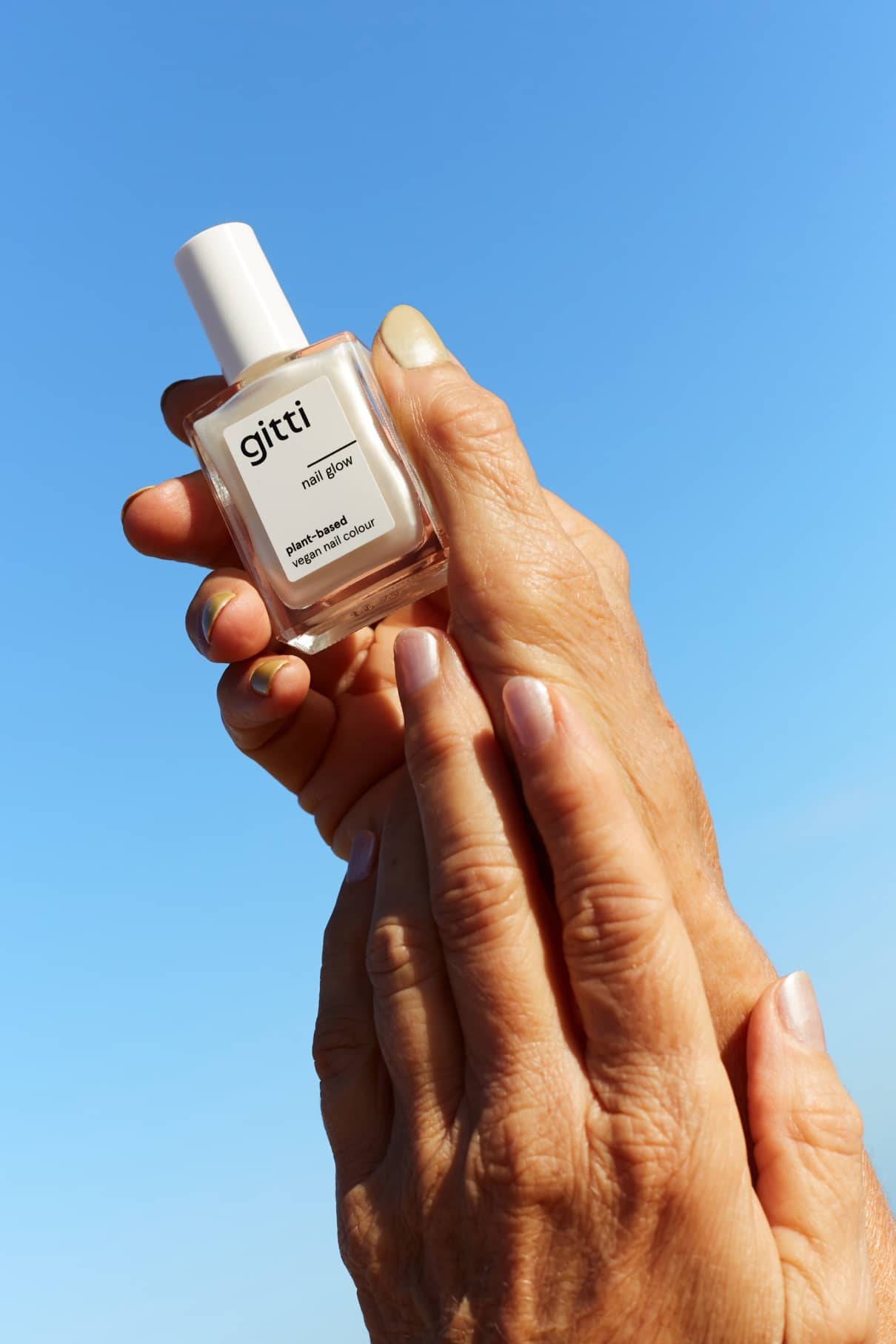 Eine Hand hält eine Flasche Nail Glow-Lack vor einem strahlend blauen Himmel, während eine andere Hand sie sanft berührt - ideal, um Ihrer Maniküre einen subtilen Chromeffekt zu verleihen.