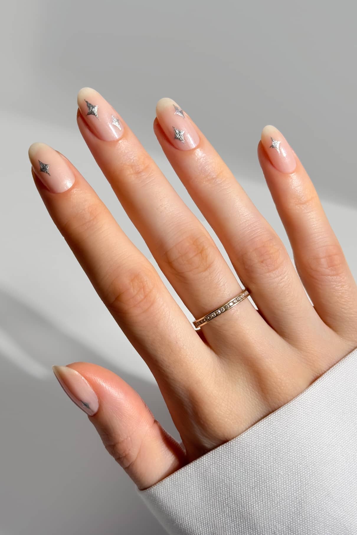 Eine Hand mit mandelförmigen Nägeln, die mit dem Nail Art Set nackt lackiert und mit kleinen silbernen Sternaufklebern verziert wurden, trägt einen einfachen goldenen Ring, fotografiert vor einem hellen Hintergrund.