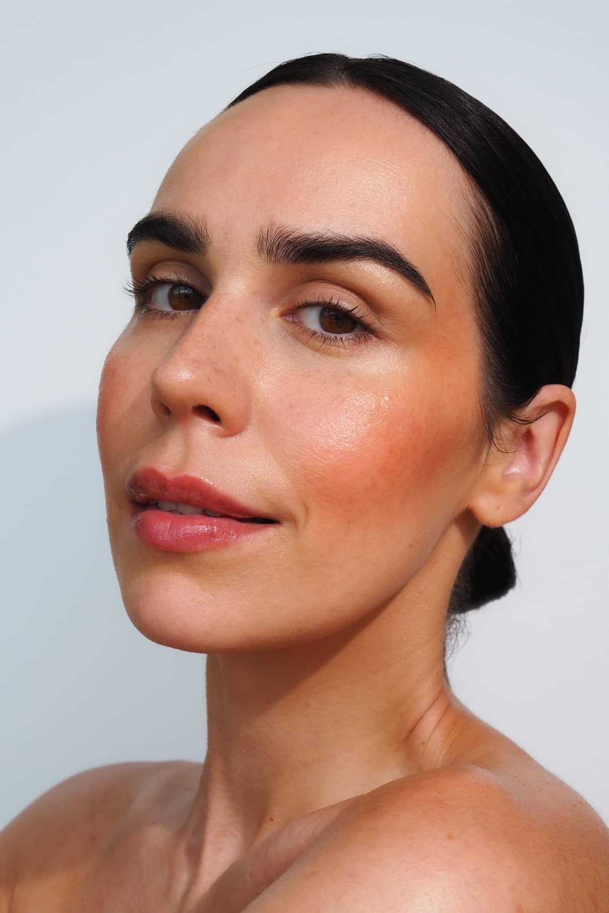 Eine Frau mit dunklem Haar posiert vor einem unifarbenen Hintergrund und trägt ein natürliches Make-up mit definierten Augenbrauen und Velvet Cheeks no. 04 für ein sonnengeküsstes Finish.