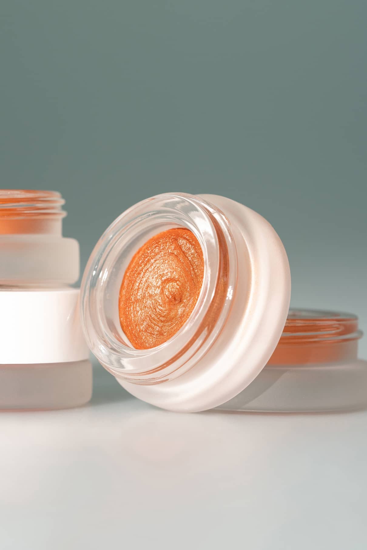 Ein offener Tiegel mit schimmerndem orangefarbenem Velvet Cheeks no. 04 Mousse-Blush steht vor seinem geschlossenen Zwilling, beide vor einem einfarbigen tealfarbenen Hintergrund - ideal für ein strahlendes, sonnengeküsstes Finish.