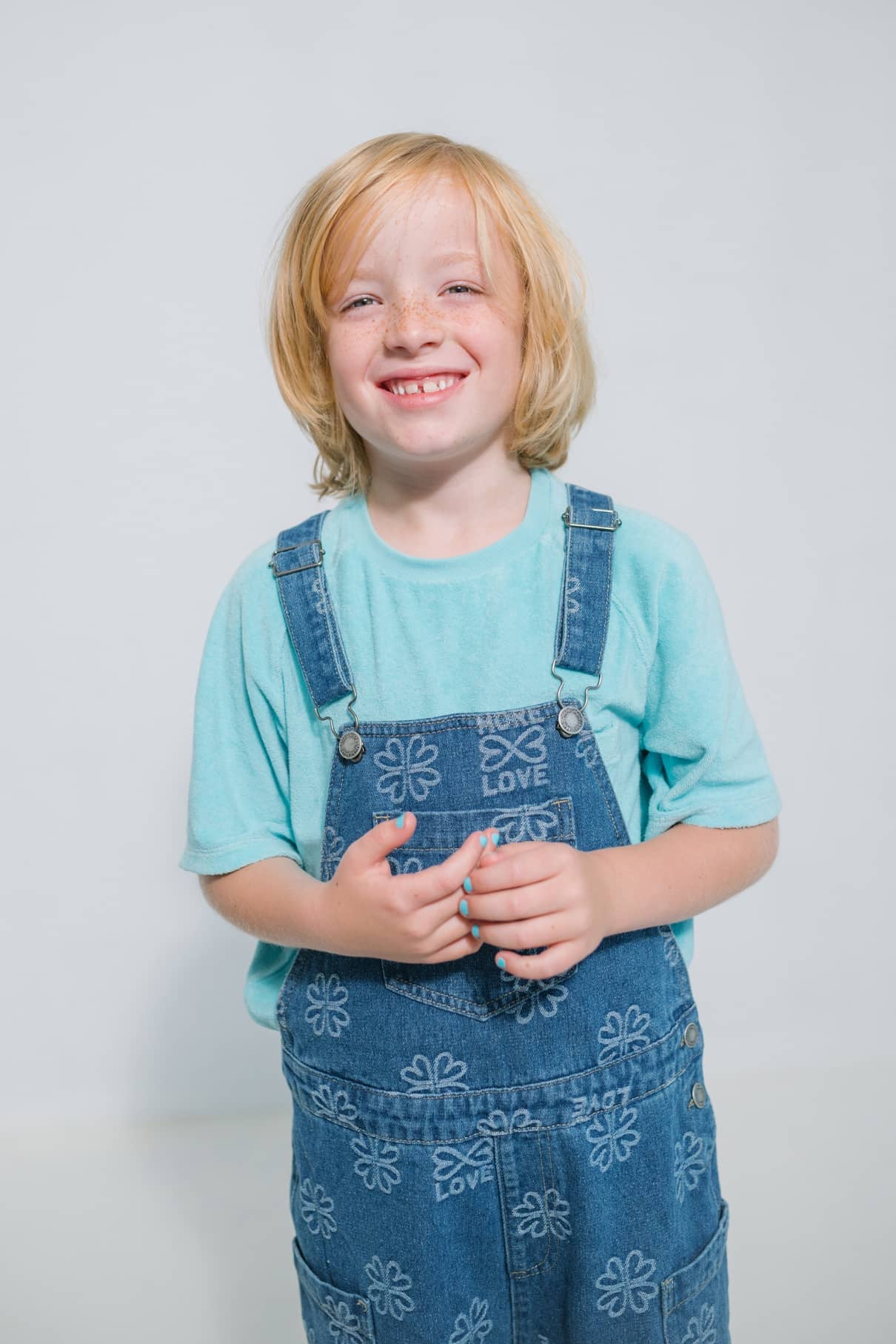 Ein lächelndes blondes Kind in einem hellblauen Hemd und einem geblümten Jeans-Overall steht vor einem schlichten Hintergrund und ist bereit, gitti kids no. 01 für einen Hauch von sicherer Kinderkosmetik.