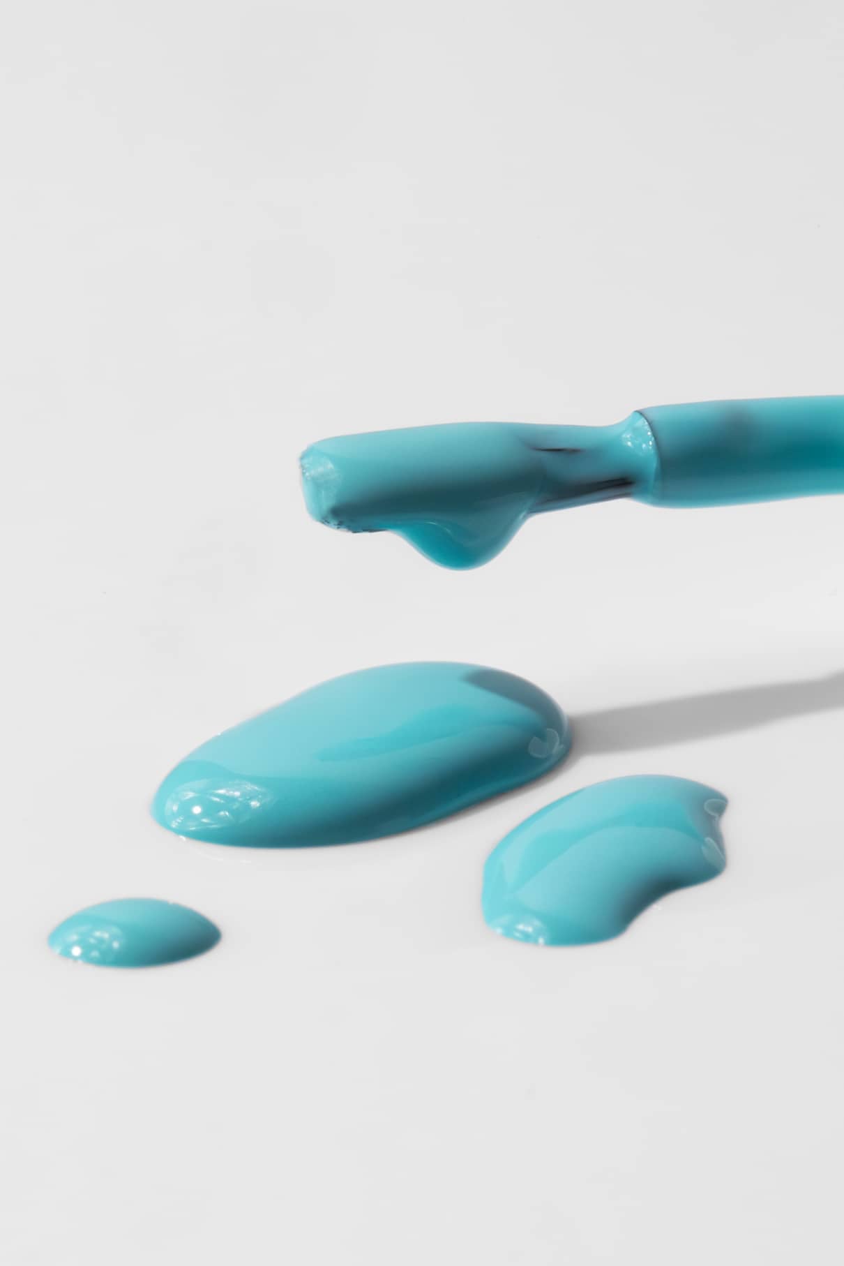 Eine Nahaufnahme eines Nagellackpinsels mit gitti kids no. 01 in Türkis, der auf eine weiße Fläche tropft und kleine Pfützen und Tropfen bildet.