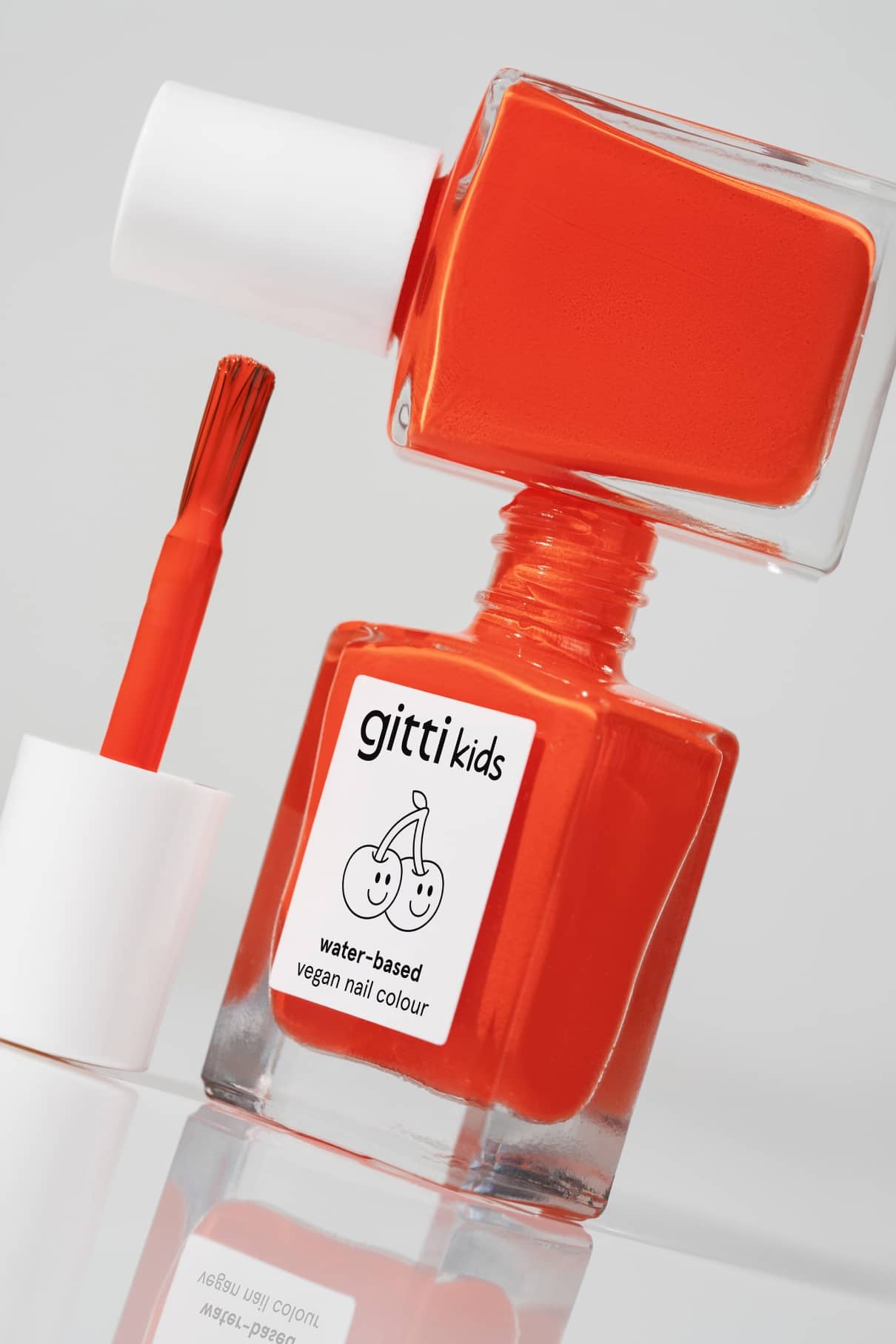 Eine Flasche der gitti kids no. 02 kinderfreundliche Nagelfarbe, leuchtend orange, wasserbasiert und vegan, steht mit dem Pinselaufsatz daneben; auf dem Etikett sind zwei Kirschen abgebildet.