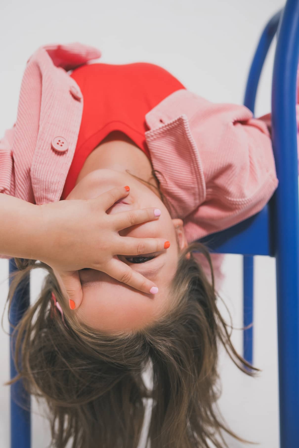 Ein Kind in rosa Jacke und rotem Hemd hängt kopfüber auf einem blauen Stuhl und verdeckt einen Teil seines Gesichts mit einer Hand, die mit gitti kids no. 02.