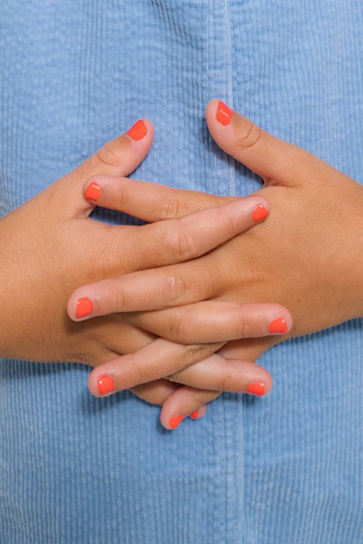 Nahaufnahme von zwei Händen mit korallenfarbenem gitti kids no. 02 Nagellack, die Finger sind ineinander verschränkt und ruhen auf einem hellblauen, strukturierten Stoffhintergrund.