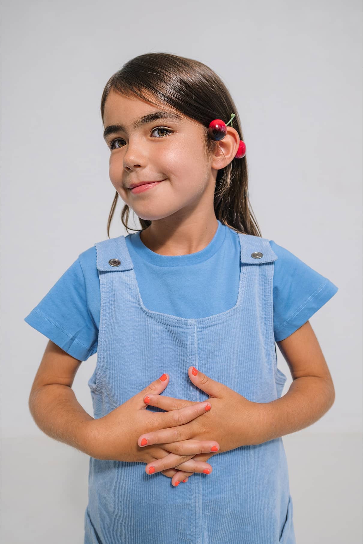 Ein junges Mädchen in blauem Hemd und Latzhose steht lächelnd mit zwei Kirschen hinter dem Ohr da - ihre Nägel sind mit gitti kids no. 02.
