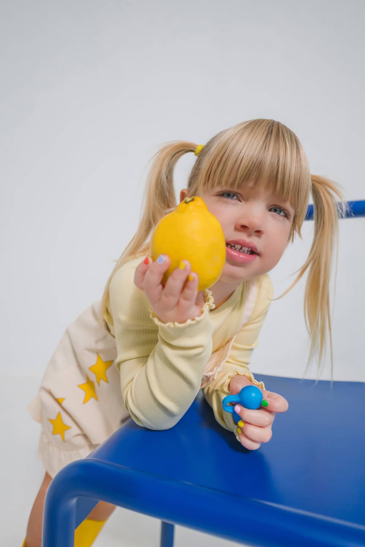 Ein junges Mädchen mit blonden Zöpfen lehnt auf einem blauen Stuhl, hält eine Zitrone und ein Spielzeug in der Hand und trägt ein helles Outfit mit gelben Sternen. Stolz zeigt sie gitti kids no. 06, ihren bunten, ungeschenkten Nagellack, auf ihren winzigen Fingernägeln.