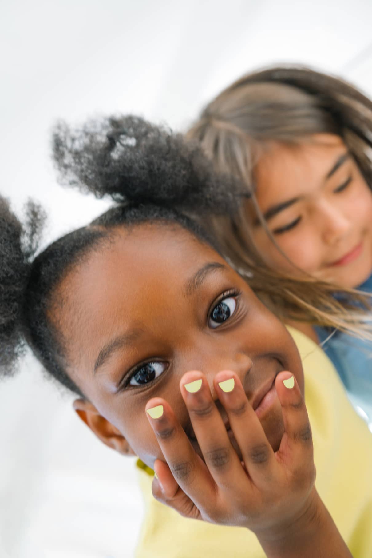 Ein Kind mit Zöpfen zeigt seine pastellfarbenen Fingernägel mit gitti kids no. 06, während ein anderes Kind mit langen Haaren im Hintergrund lächelt.