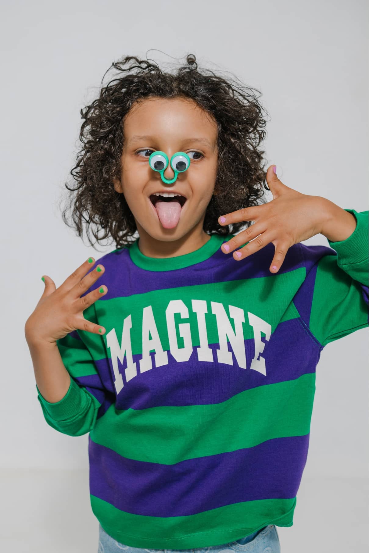 Ein Kind in einem grün-lila "IMAGINE"-Shirt hebt die Hände, streckt die Zunge heraus und zeigt stolz den gitti kids no. 07 Nagellack, während es Spielzeugringe auf der Nase balanciert.