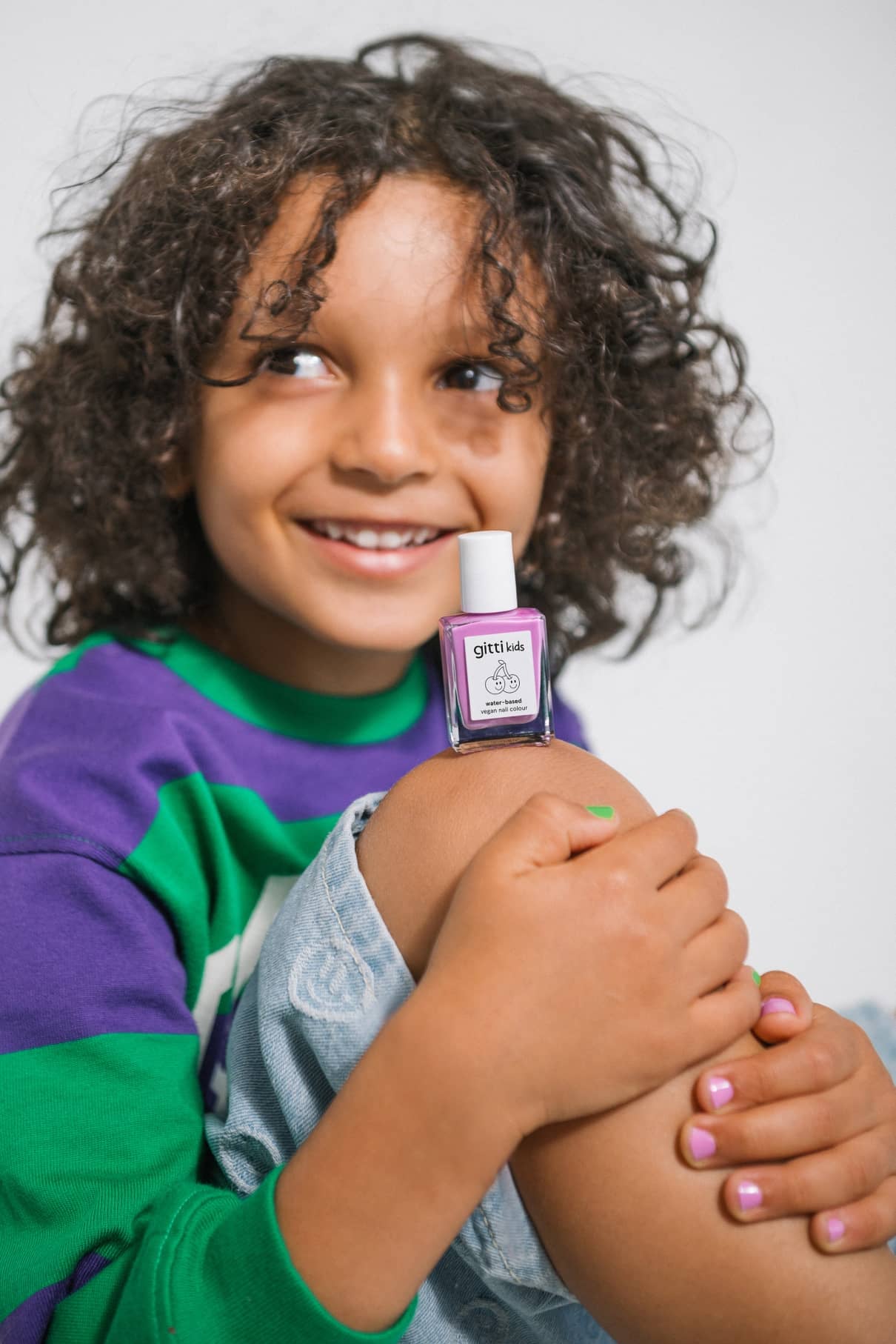 Ein lächelndes Kind mit lockigem Haar, in einem lila-grünen Pullover, hält sich das Knie und zeigt gitti kids no. 07-eine kinderfreundliche Nagelfarbe-auf ihren schön lackierten Nägeln.
