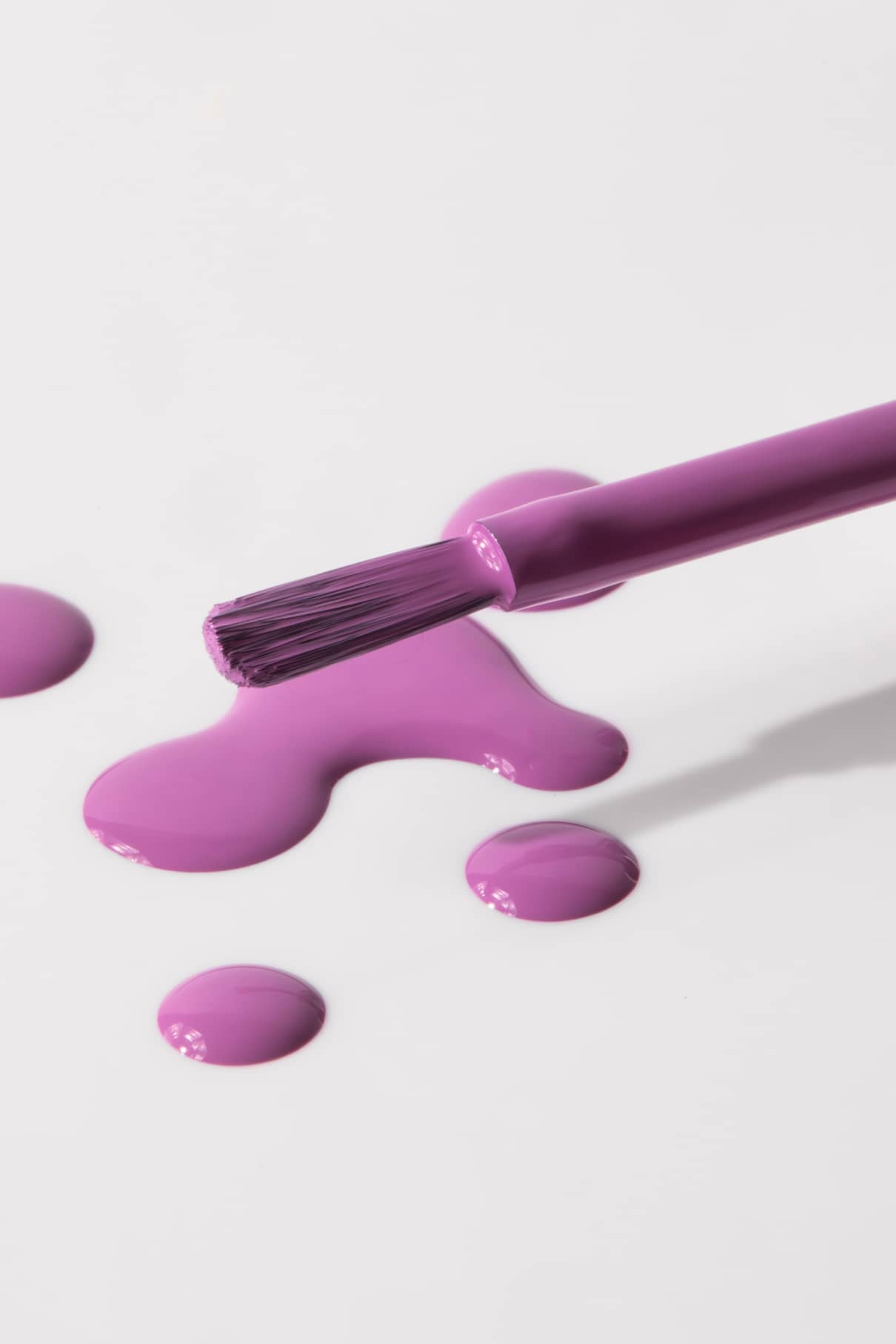 Ein in gitti kids no. 07 lila Nagellack getaucht ist, ruht auf einer weißen Fläche, umgeben von kleinen Pfützen und Tropfen in demselben leuchtenden Farbton.