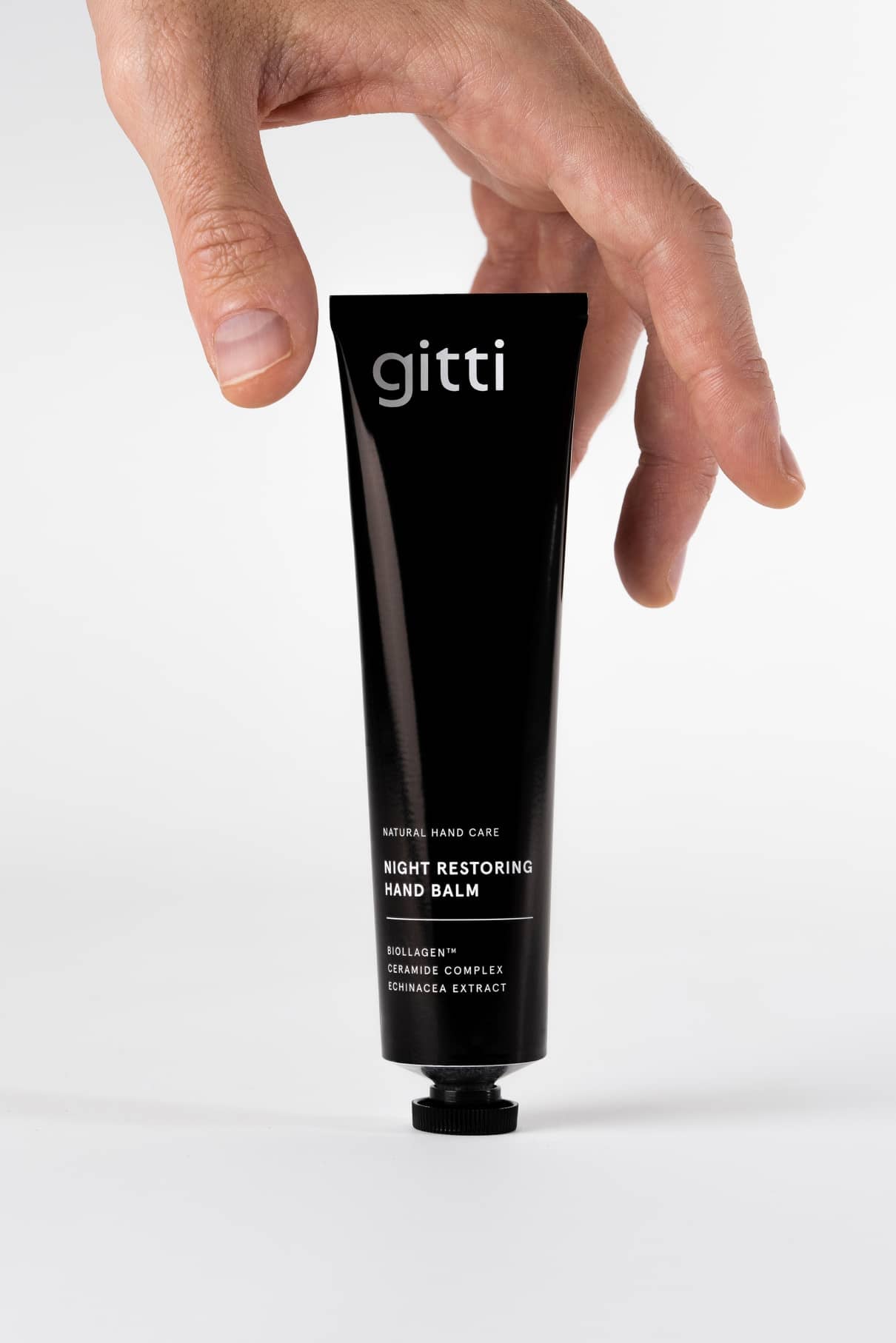 Eine Hand greift nach einer schwarzen Tube 24 Hour Hand Therapy, die glatte Hände als Teil Ihrer 24h-Pflege-Routine verspricht, und steht aufrecht auf weißem Hintergrund.